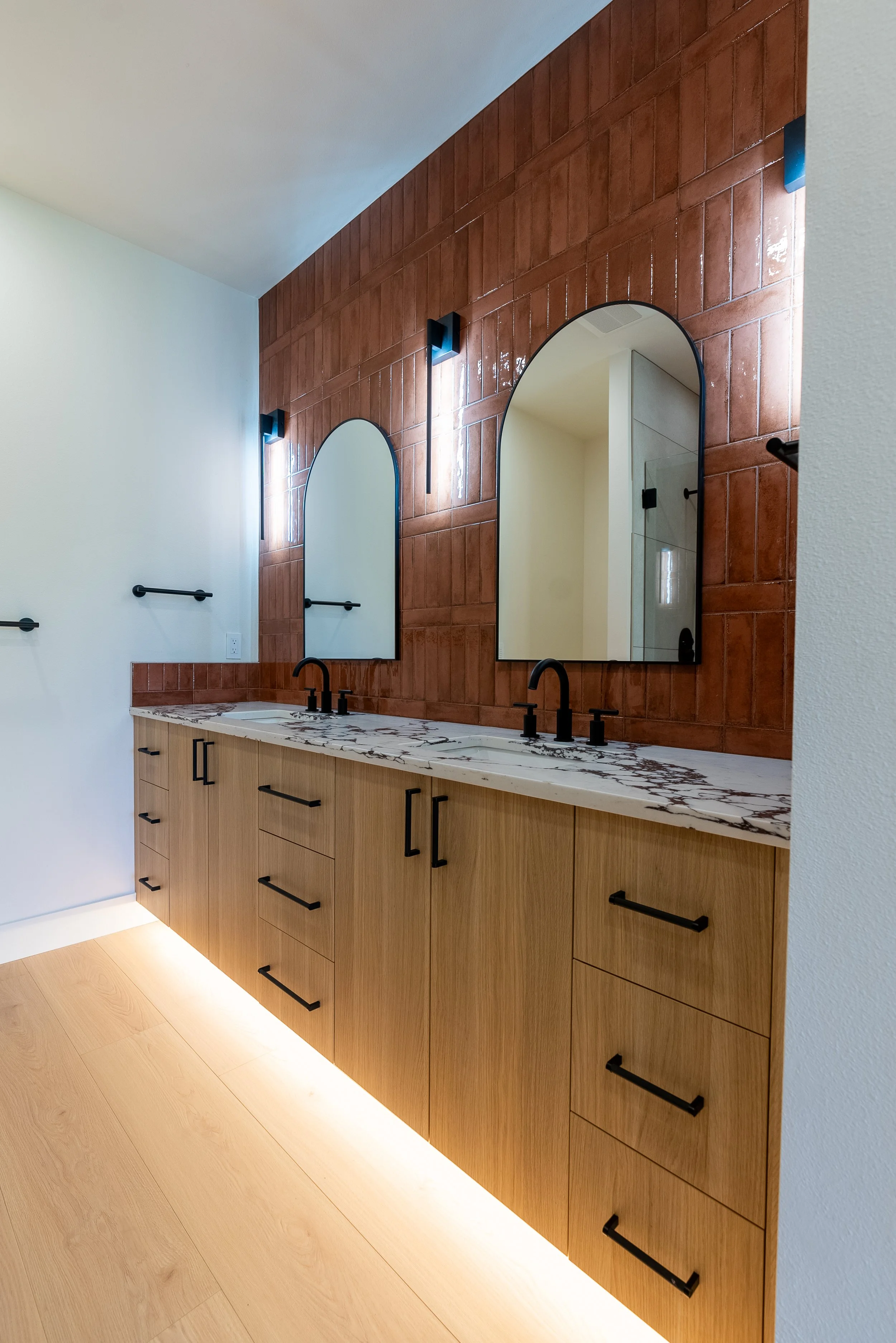 Modern bathroom vanity with light wooden cabinetry, black hardware, a marble countertop, and two mirrors mounted on a terracotta tile wall.