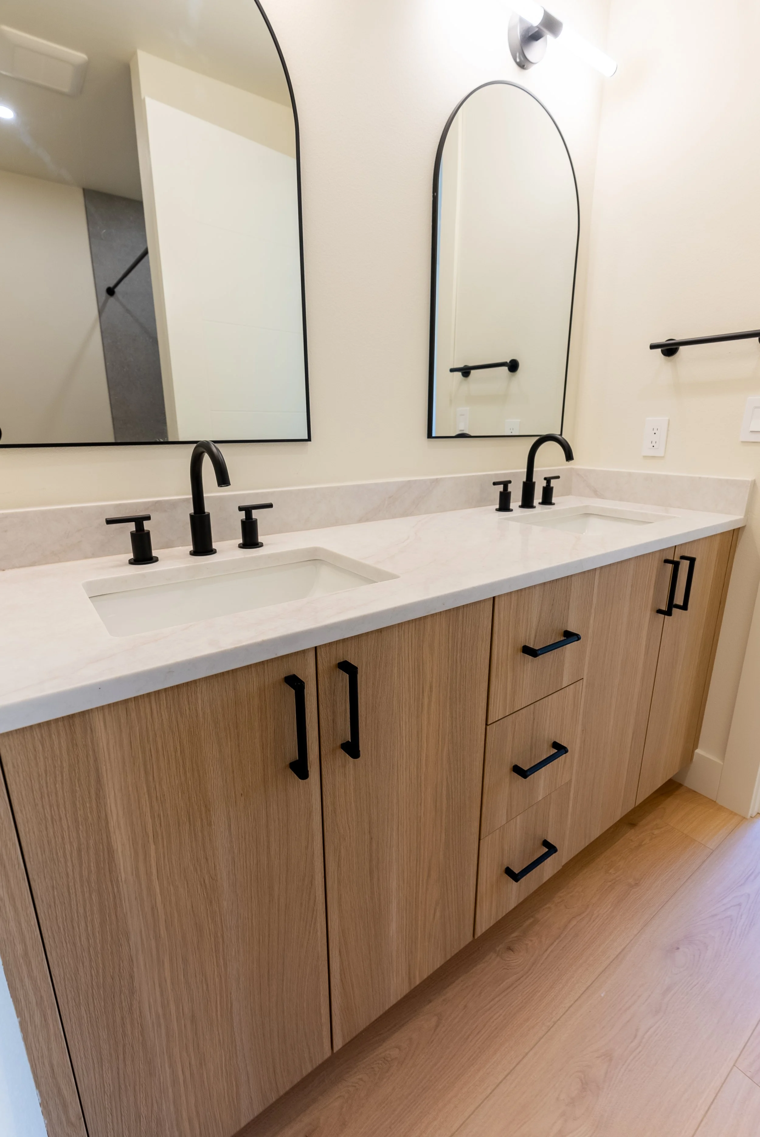 Bathroom vanity with two sinks, black matte faucets, wood cabinets, large arched mirrors, black hardware, light-colored countertop, wall-mounted light fixture.