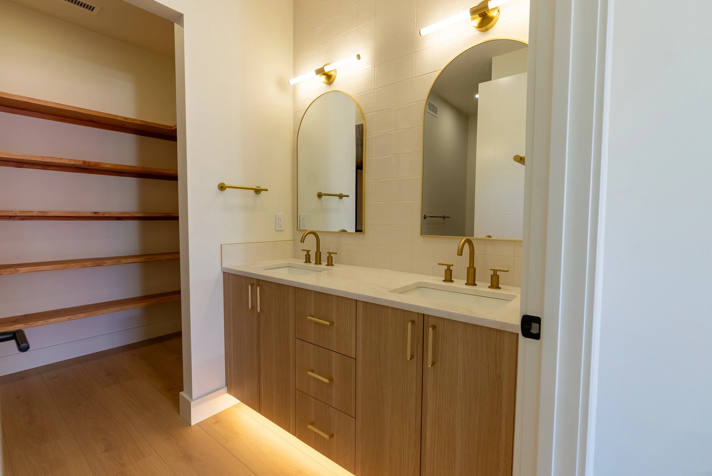 Modern bathroom with two arched mirrors, gold fixtures, wooden vanity, and open shelving