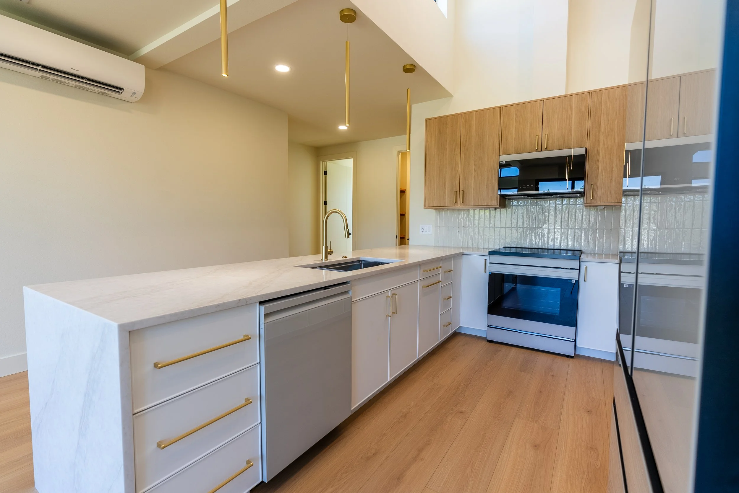 Modern kitchen with white island, light wood cabinets, black oven, microwave, and gold accents, hardwood floors, and a white wall with air conditioning unit.