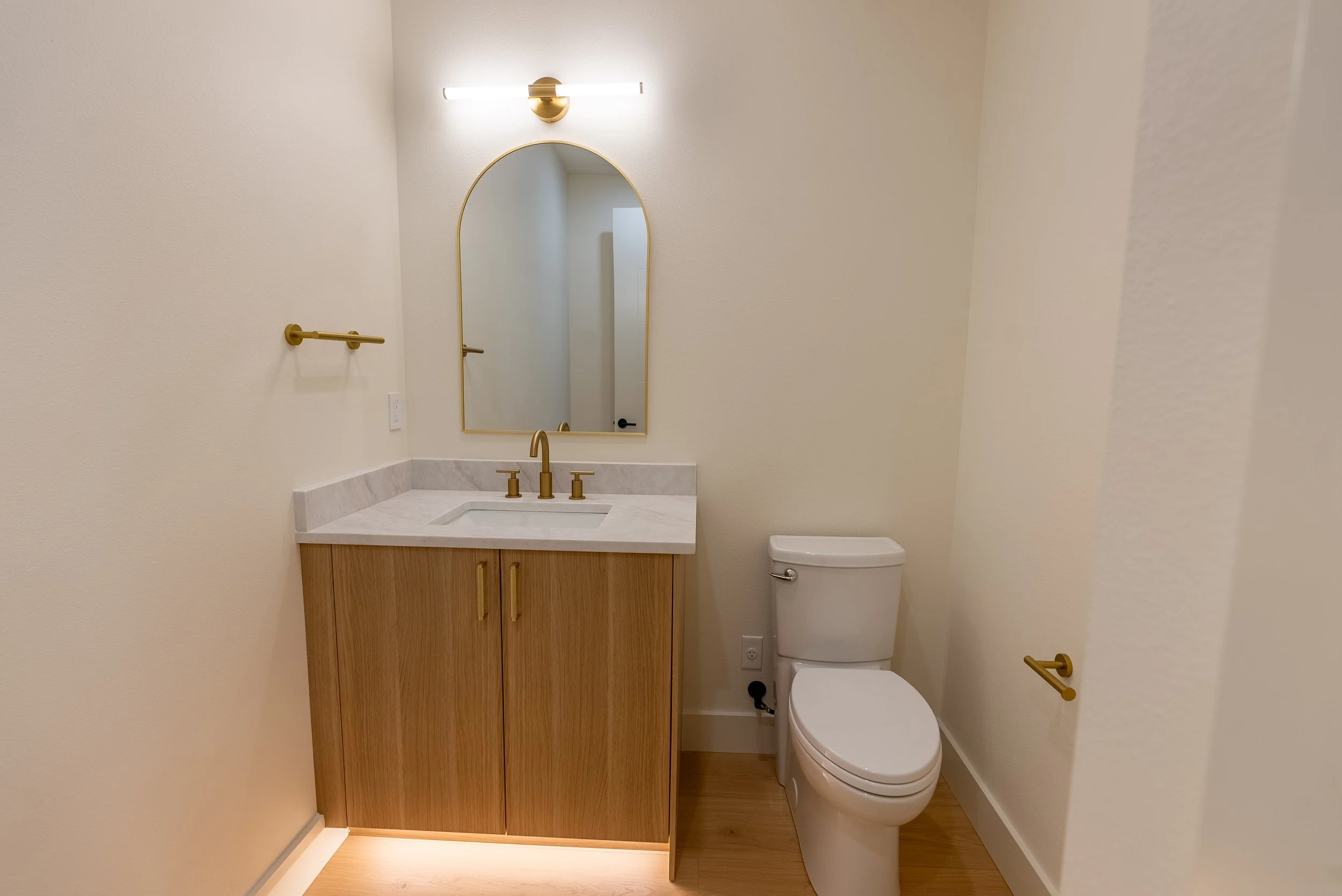 A small bathroom with a wood cabinet vanity, a white marble countertop, a gold faucet, a tall mirror, gold towel and toilet paper holders, and a white toilet. The walls are painted off-white, and the flooring is wood. There is a modern wall-mounted l
