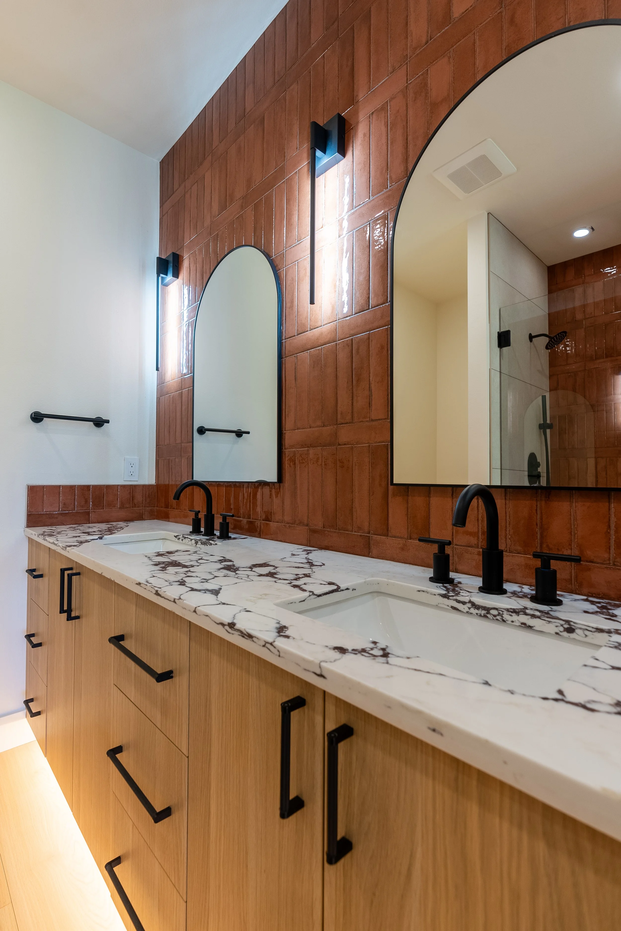Modern bathroom vanity with a marble countertop, black faucets, wooden cabinets, and a large mirror with black trim. The wall is tiled with terracotta-colored tiles, and there are black wall sconces and a towel bar.