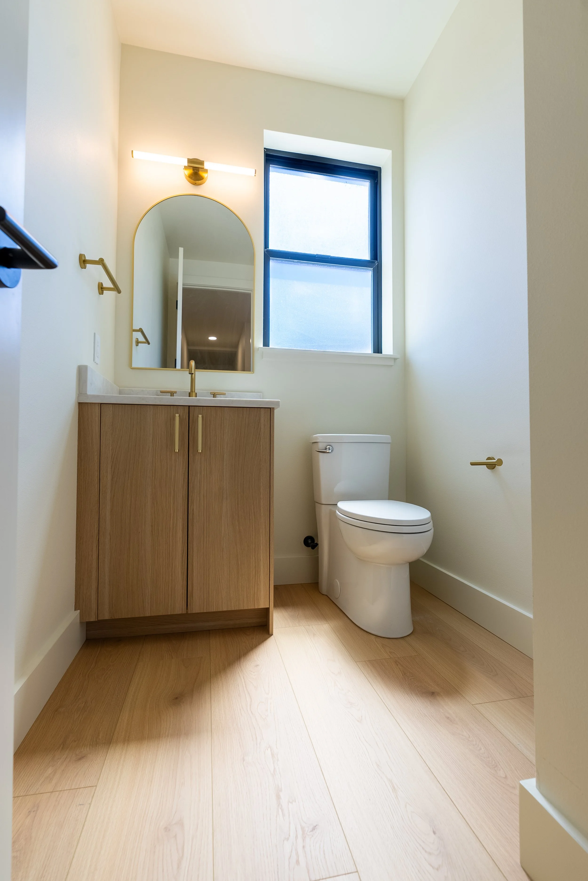 A modern bathroom with light wood flooring, a white toilet, a wooden vanity with a white countertop, and a large window with frosted glass. There is a wall-mounted mirror above the vanity, a gold-tone wall light fixture, and gold fixtures on the vani