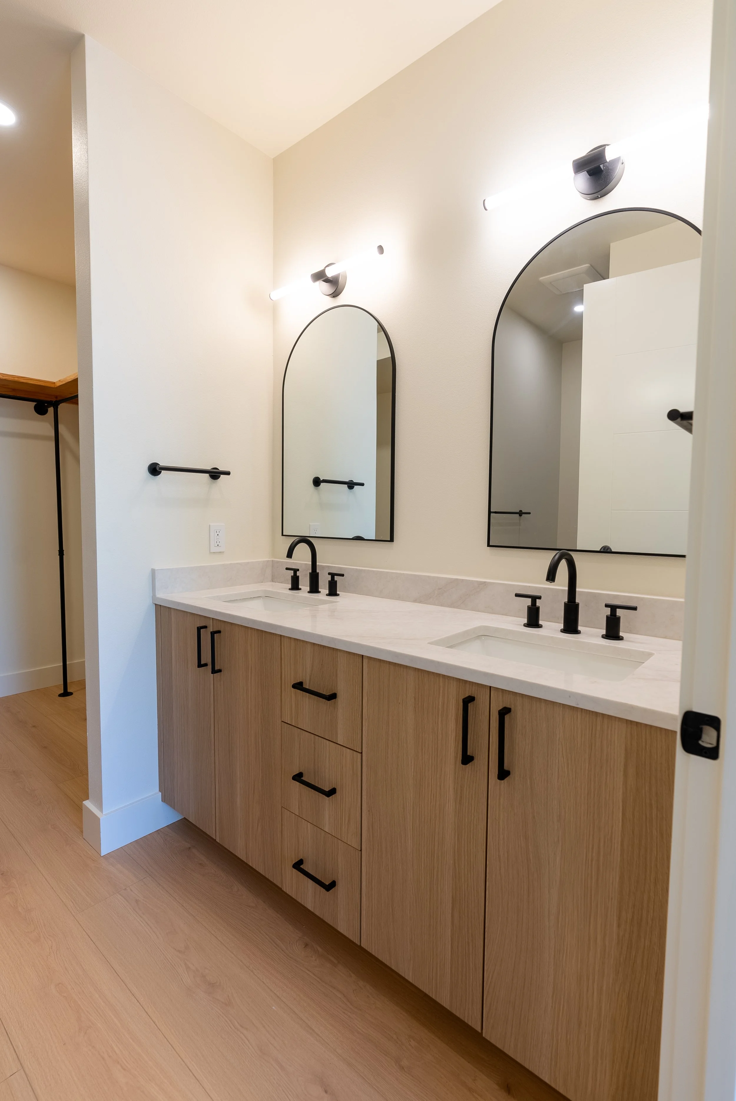 Modern bathroom vanity with two oval mirrors, black faucet, light wood cabinets, and black hardware. There are two light fixtures above each mirror. The countertop is white with a built-in sink.