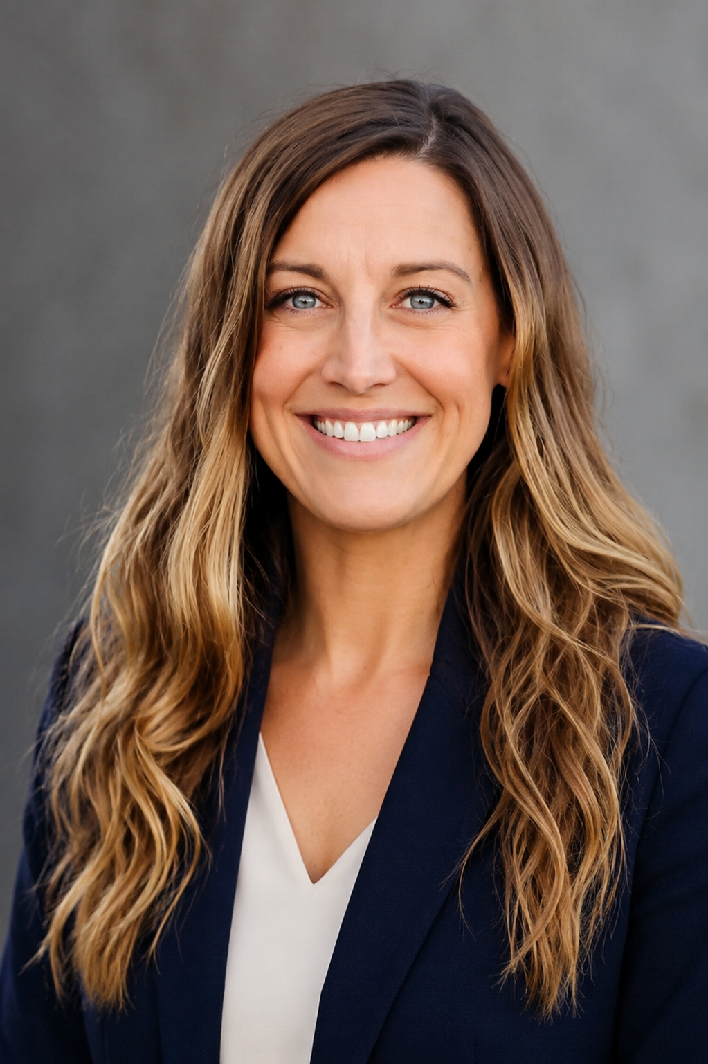 Headshot of a smiling woman with long wavy light brown hair wearing a dark blazer over a white top, standing against a plain gray background.