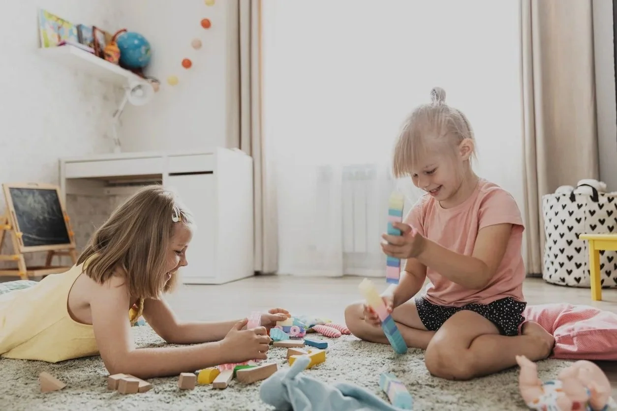 Two young girls playing with colorful toys and building blocks on a carpet in a bright room with a window, curtains, and some furniture, working on improving communication skills during in-home speech therapy for kids in Appleton, WI.