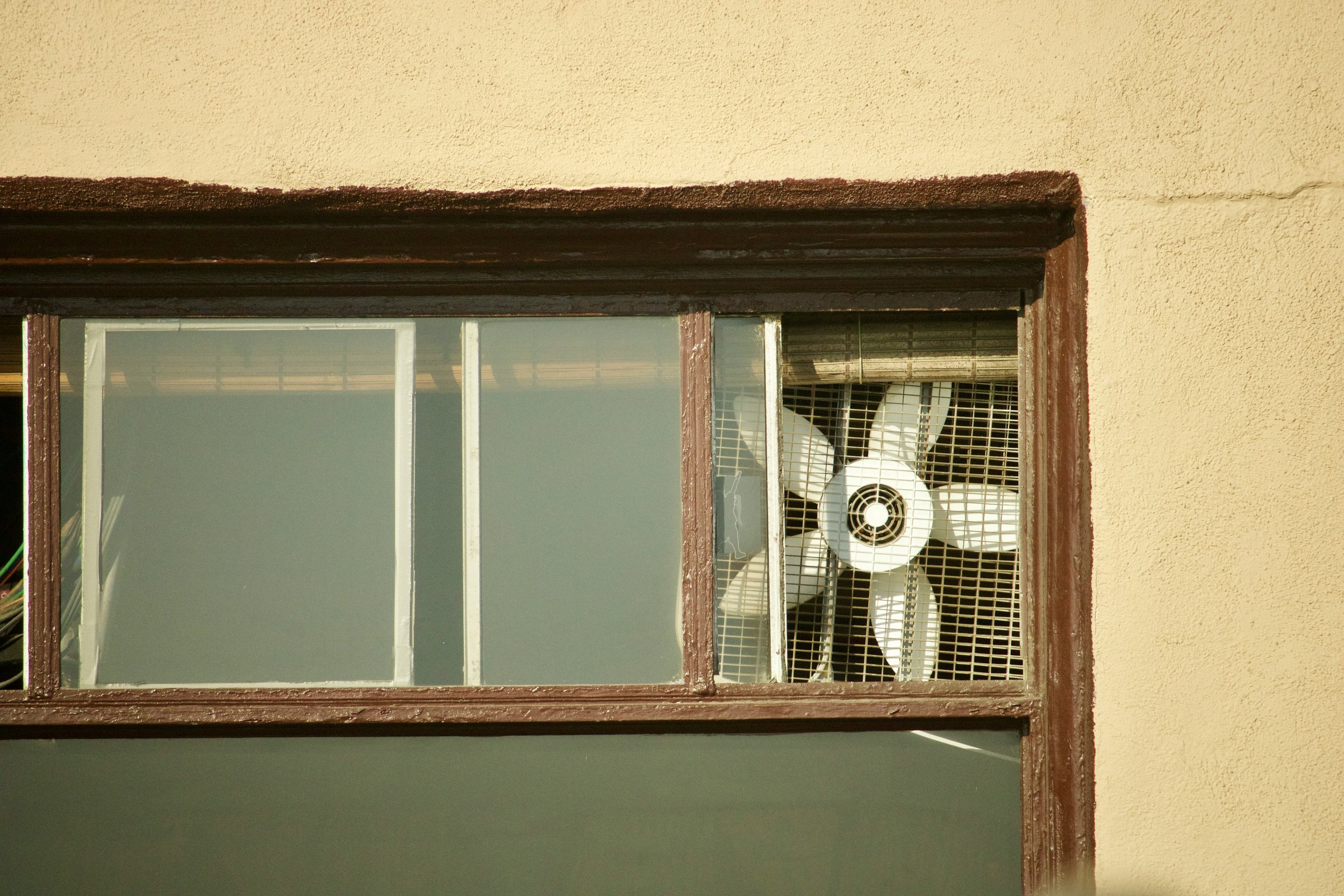 Close-up of a window with a white fan behind the grid and a yellow wall.
