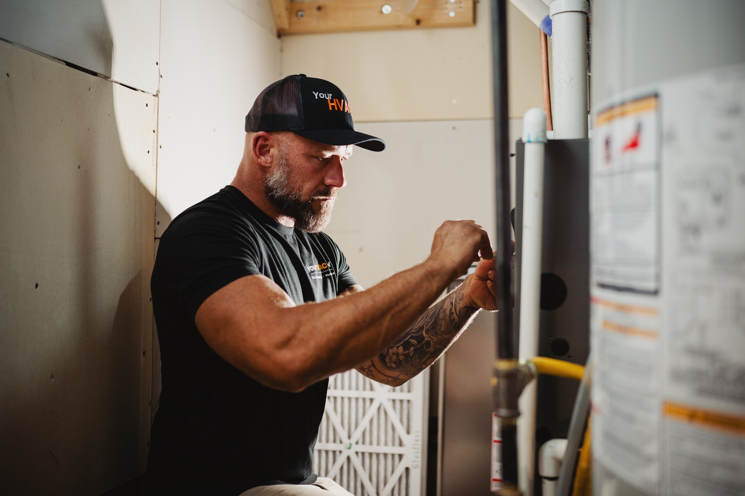 A man with a gray beard wearing a black cap and black T-shirt working on a heating unit in a residential furnace room.