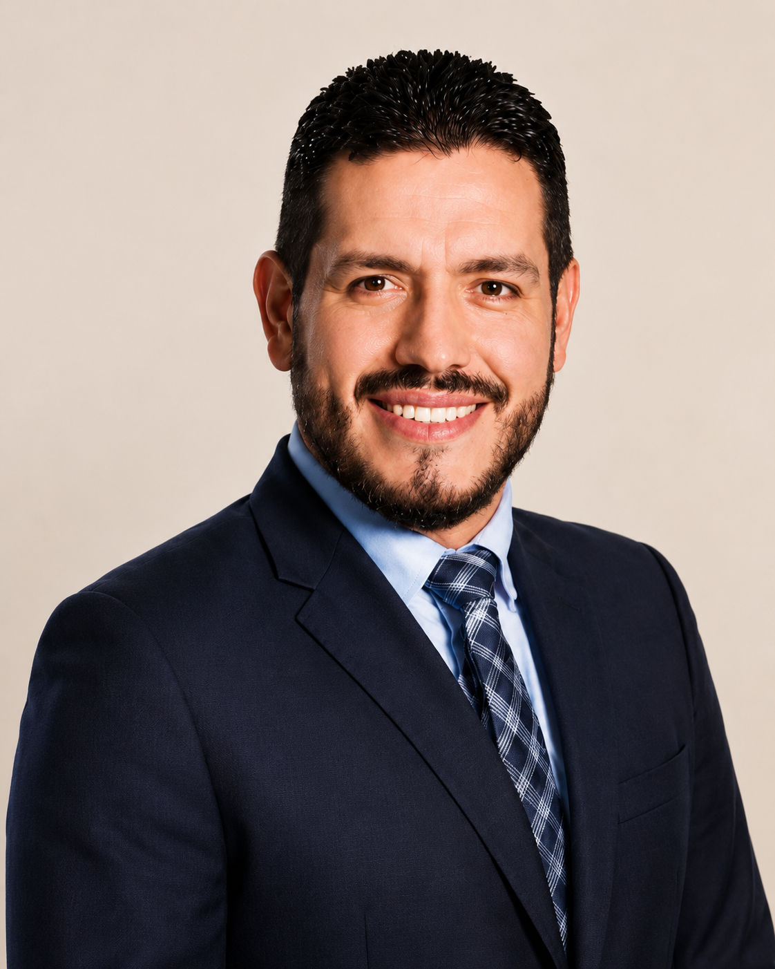 Retrato de un hombre con barba, pelo oscuro y sonrisa, vestido con saco azul, camisa clara y corbata a cuadros, contra fondo beige.