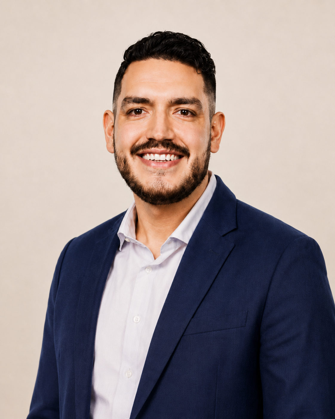 Retrato de un hombre con barba y cabello oscuro, vestido con saco azul y camisa blanca, sonriendo y posando frente a fondo beige