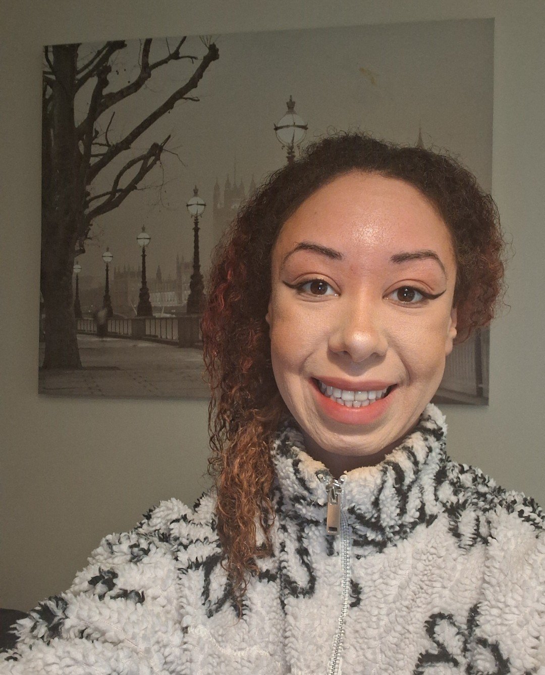 A woman smiling in front of a black and white cityscape wall art. She has curly brown hair with reddish highlights and is wearing a cozy white and black patterned fleece jacket.
