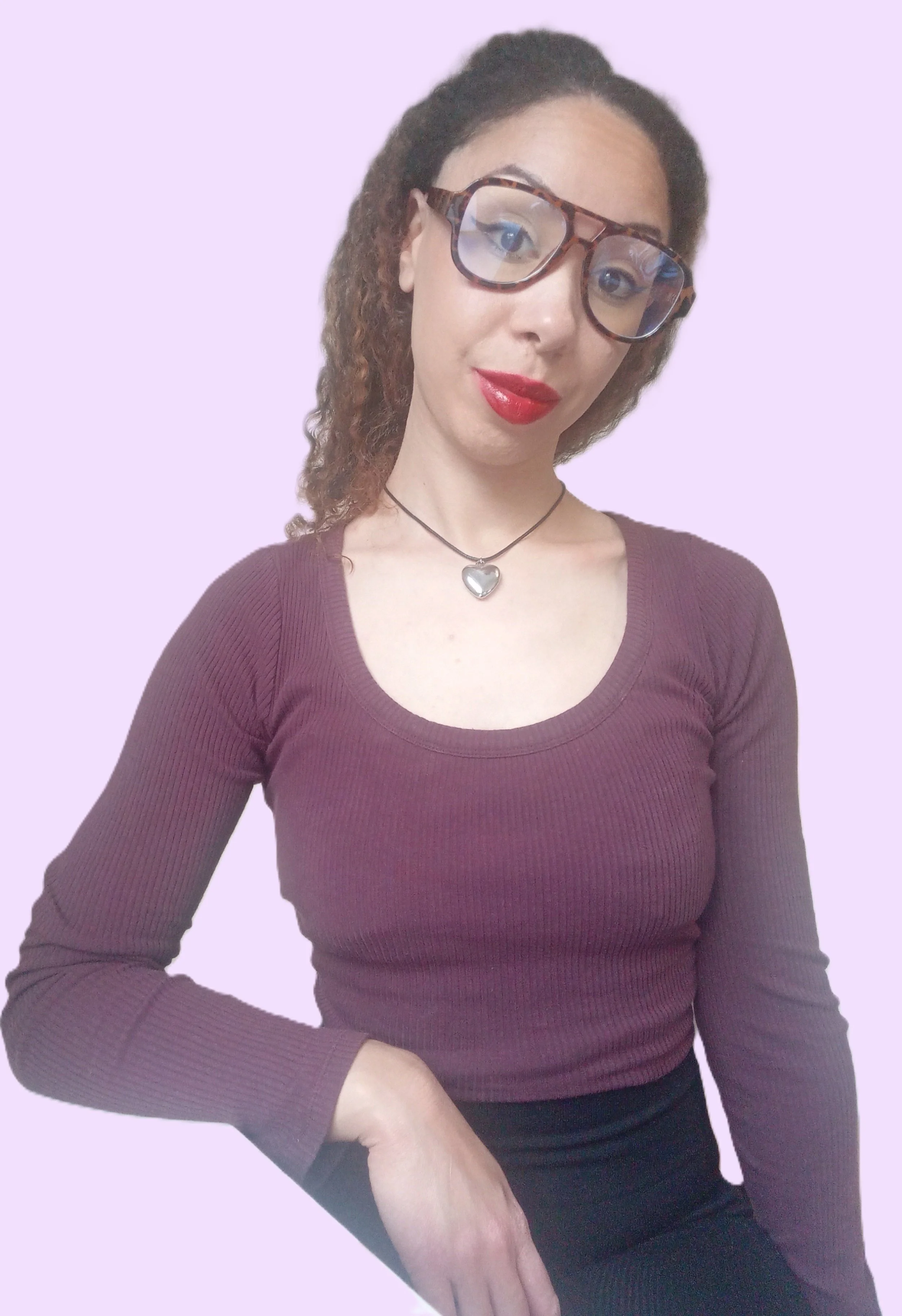 A woman with curly brown hair, wearing glasses with a tortoise shell pattern, a heart-shaped necklace, a maroon long-sleeve top, and red lipstick, against a light purple background.