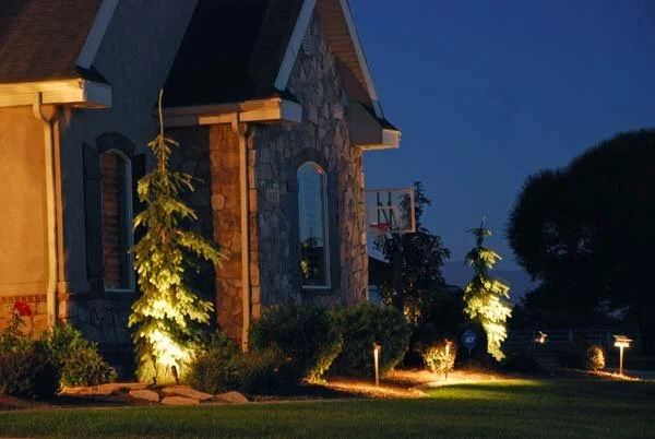 A stone house with a blue roof illuminated by outdoor lighting at night. There are small lit trees and bushes in the yard, and a basketball hoop is attached to the house.