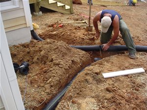 A person working on installing a drainage pipe outside a house, digging in the dirt.