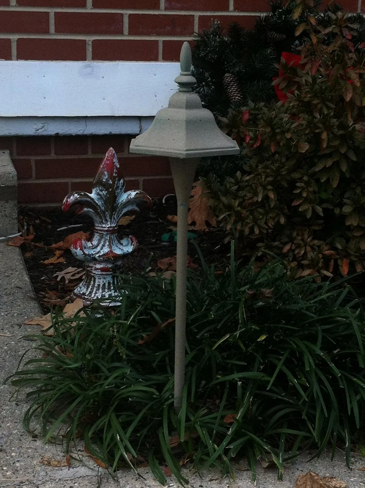 A garden scene with a decorative metallic ornament resembling a fleur-de-lis, a garden light post, and various plants including a bush with red and green leaves.