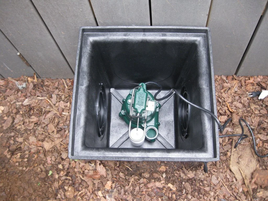 An outdoor grow box with a green grow light in the center, two black fans on either side, and an electrical cord connected to the device, placed on dirt with mulch in front of a wooden fence.
