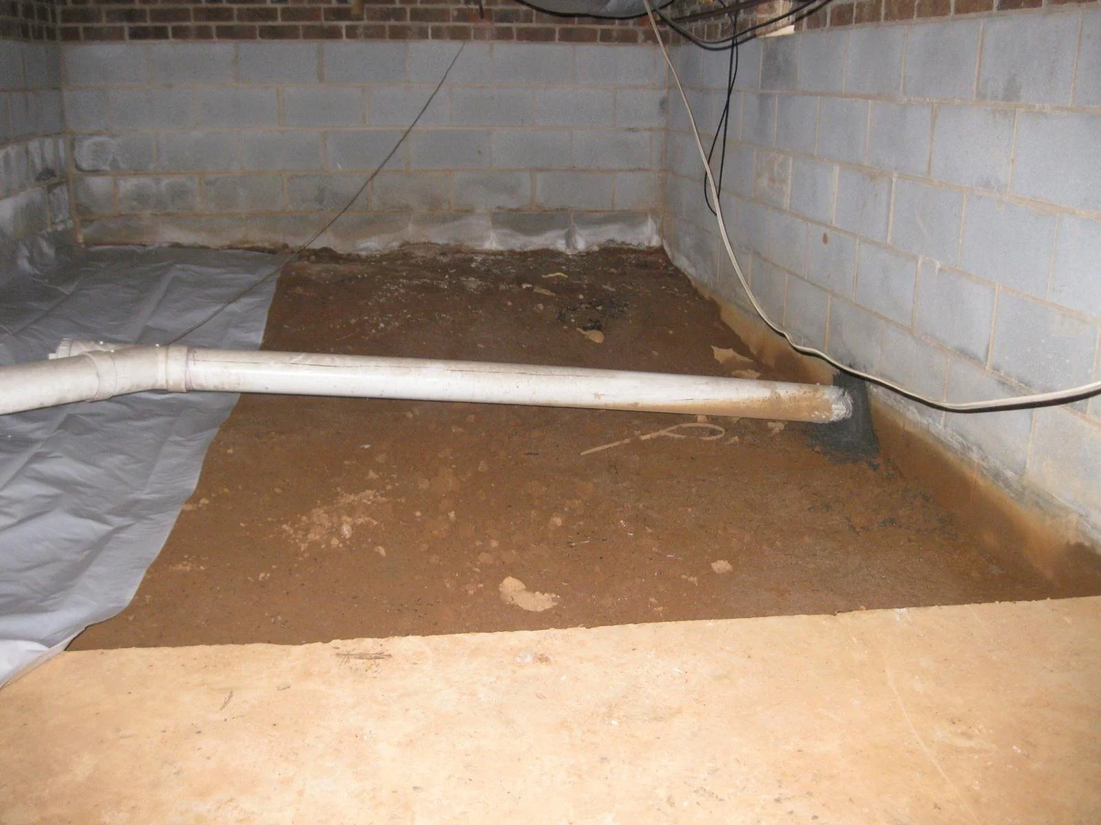 A construction or renovation site showing a section of a basement with exposed soil, a white PVC pipe, electrical wires, concrete block walls, and a partially installed vapor barrier or plastic sheet on the ground.