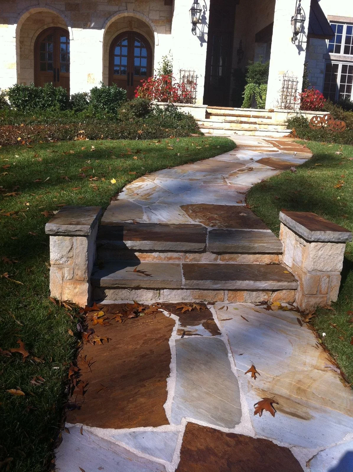 Stone pathway with stone steps leading to a house with arched wooden doors and lantern lights, bordered by bushes and fall leaves.