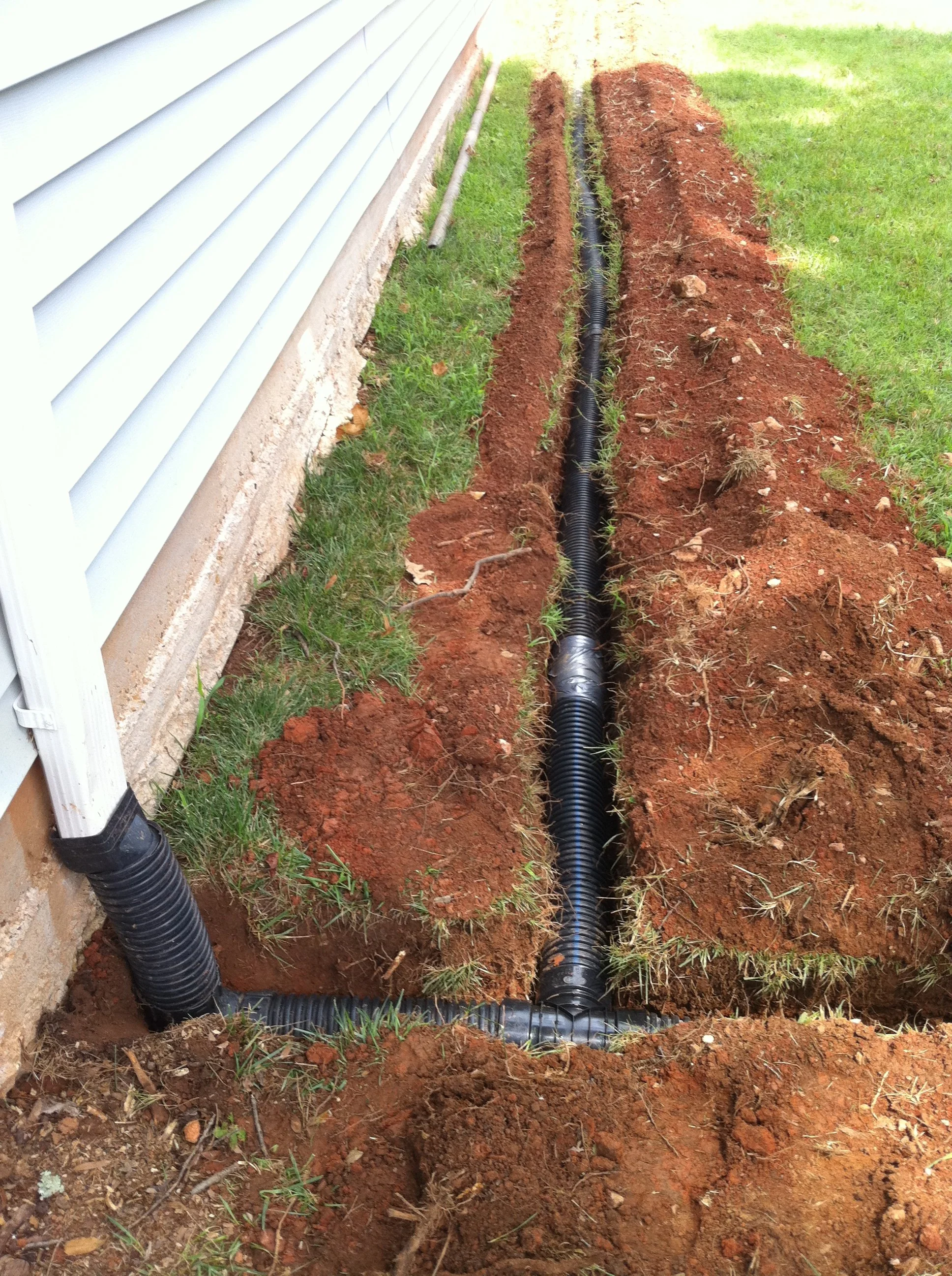 A trench dug in the ground next to a house with grey siding, revealing black corrugated drainage pipes.