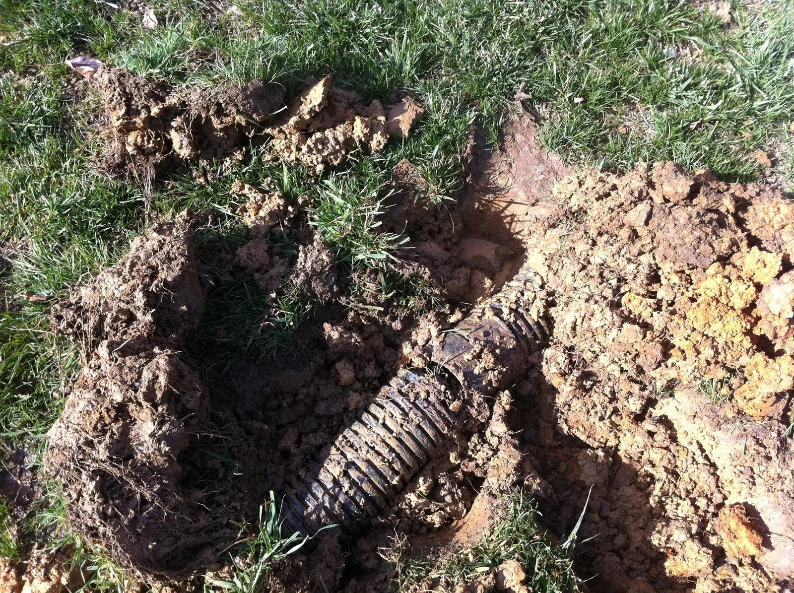 An exposed section of earth showing a partially buried, rusted pipe with a corrugated surface, surrounded by green grass and disturbed soil.