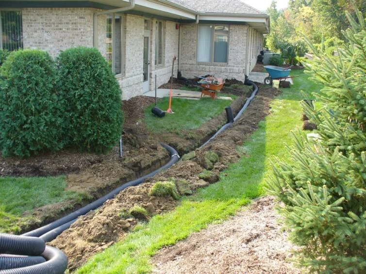 Garden bed construction with black drainage piping, tools, and supplies near a brick house with bushes and a grassy yard.