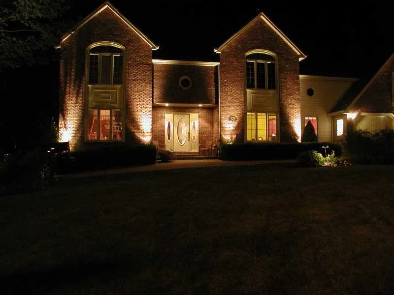 A large brick house illuminated at night with exterior lighting highlighting its architectural features, including tall windows and a central front door.