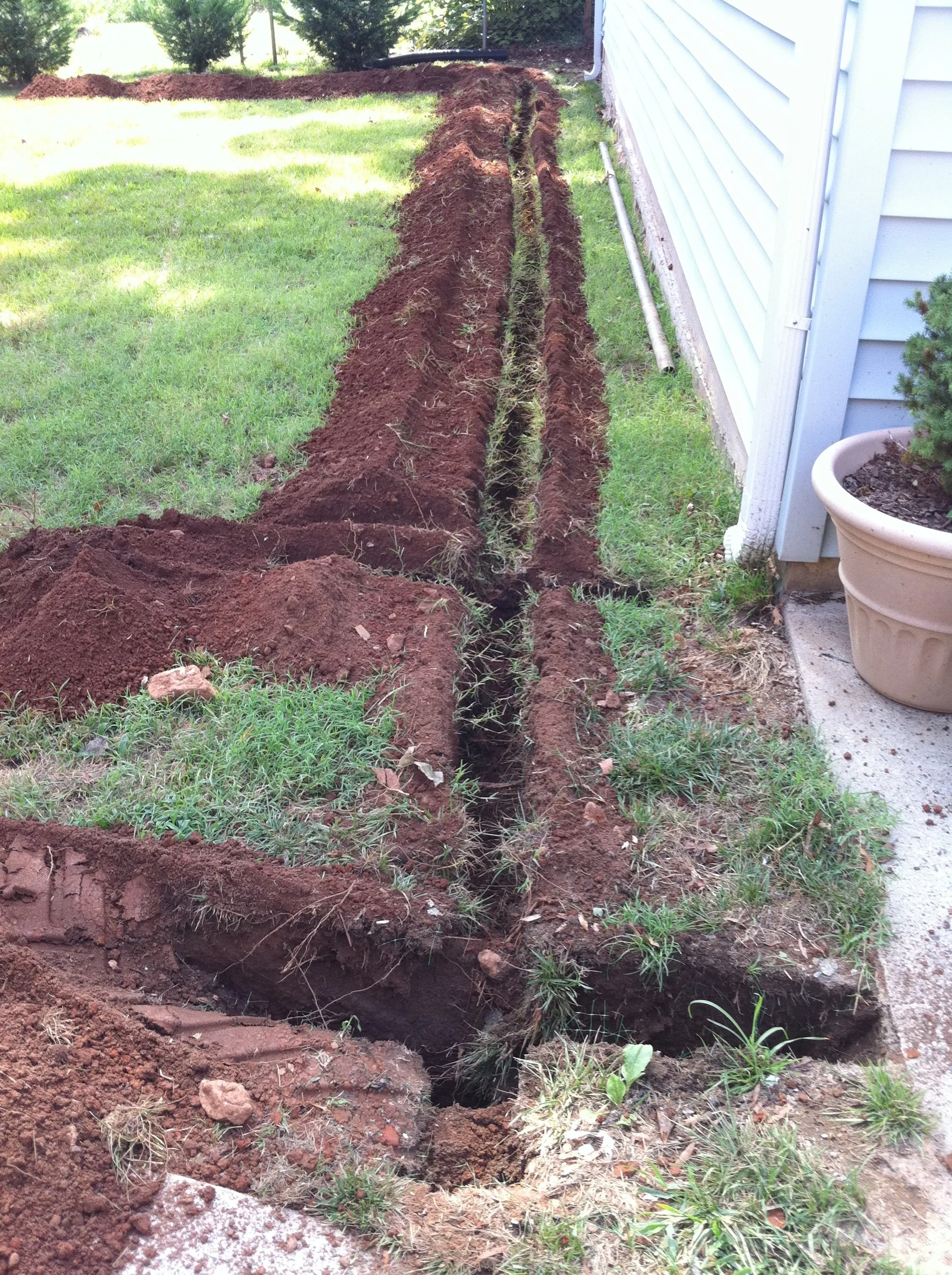 A garden trench dug beside a house, with dark soil and some grass and plants nearby.