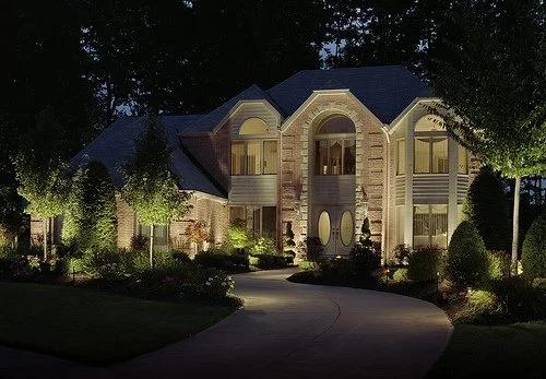 Large illuminated house at night with a curved driveway, trees, and landscaped yard.