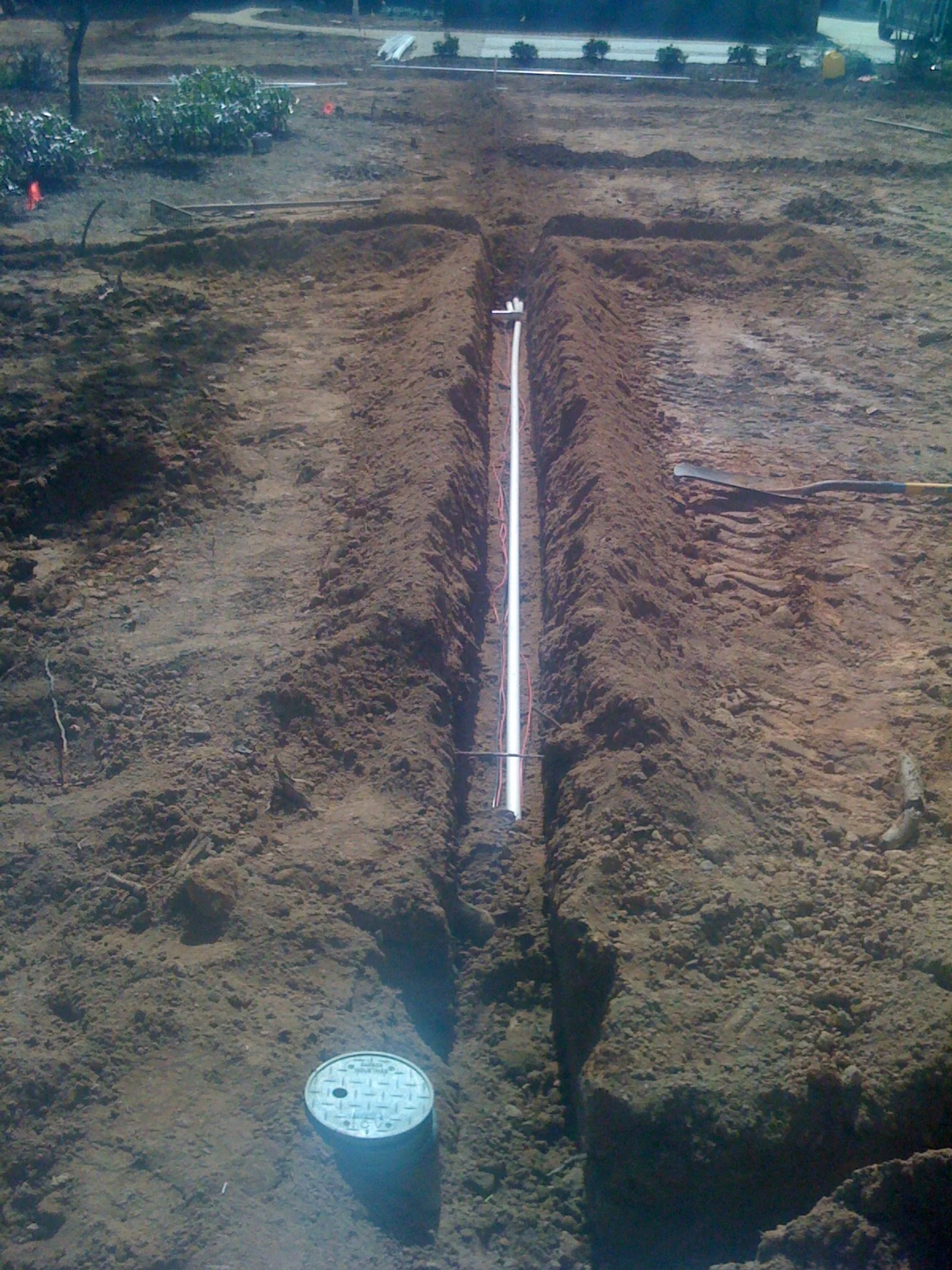 A trench in the ground with electrical wiring and a white pipe running through the middle. A manhole cover is visible in the foreground, and there are tools and soil around the trench.