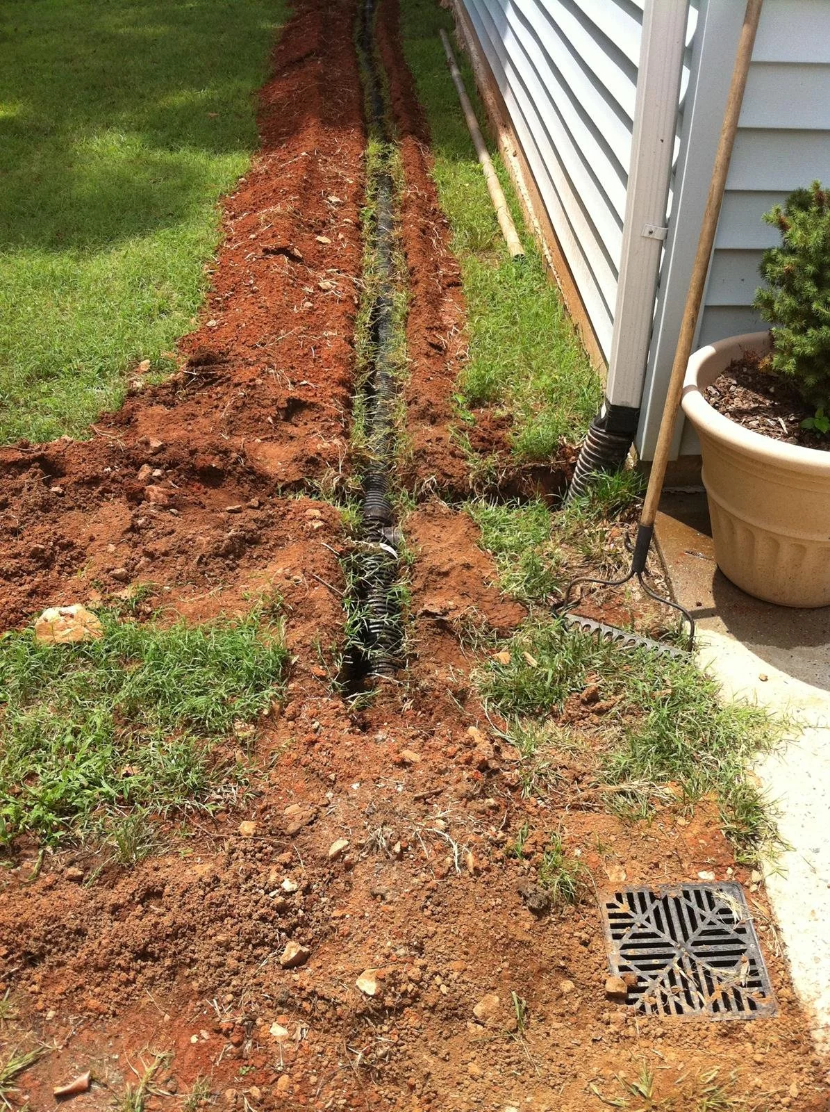 Soil trench alongside a house with a black drainage pipe and a drain cover, grass and potted plant nearby.