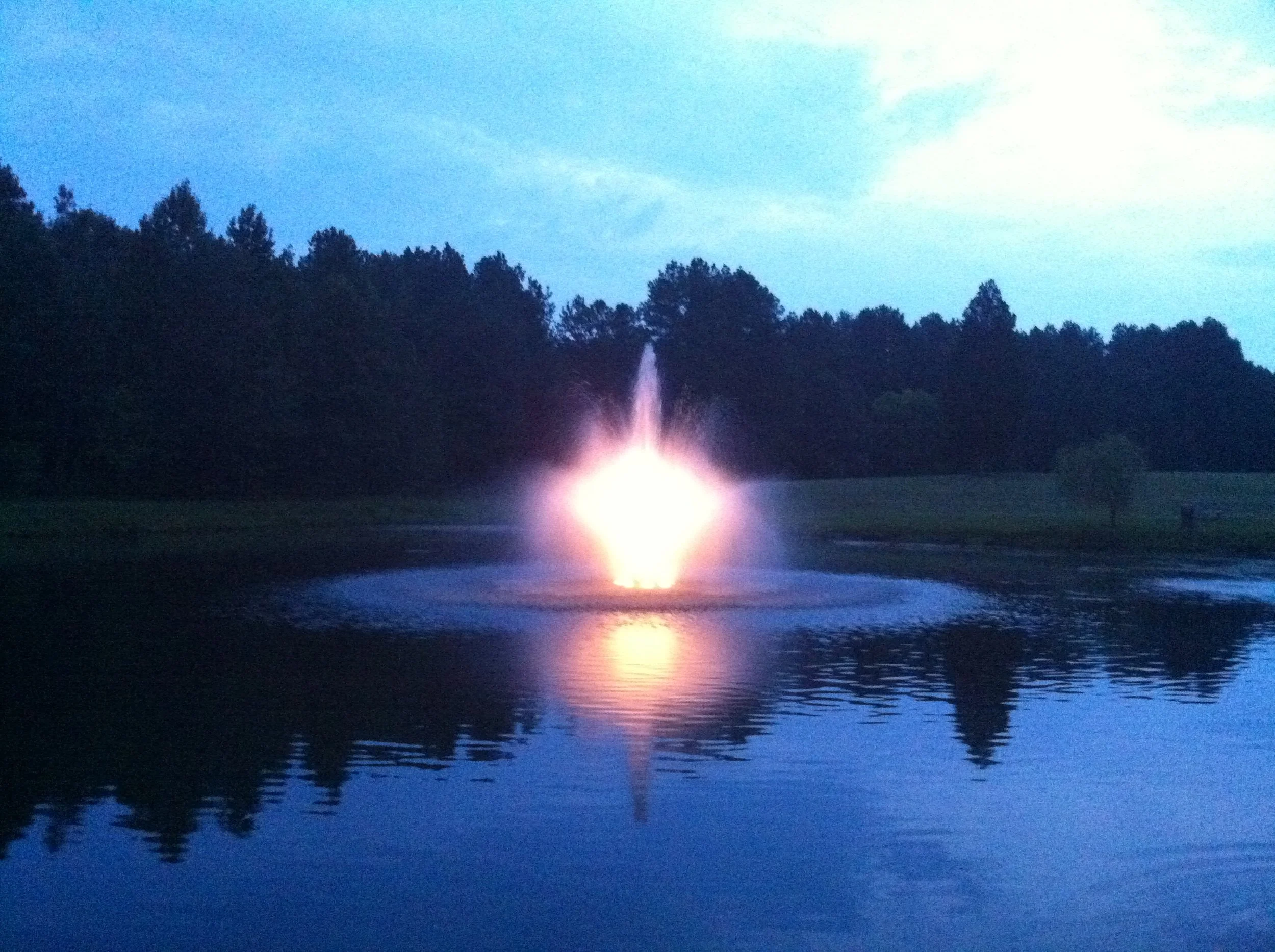 A fountain shooting water into the air at dusk or night, with a reflection on a calm body of water and a silhouette of trees in the background.