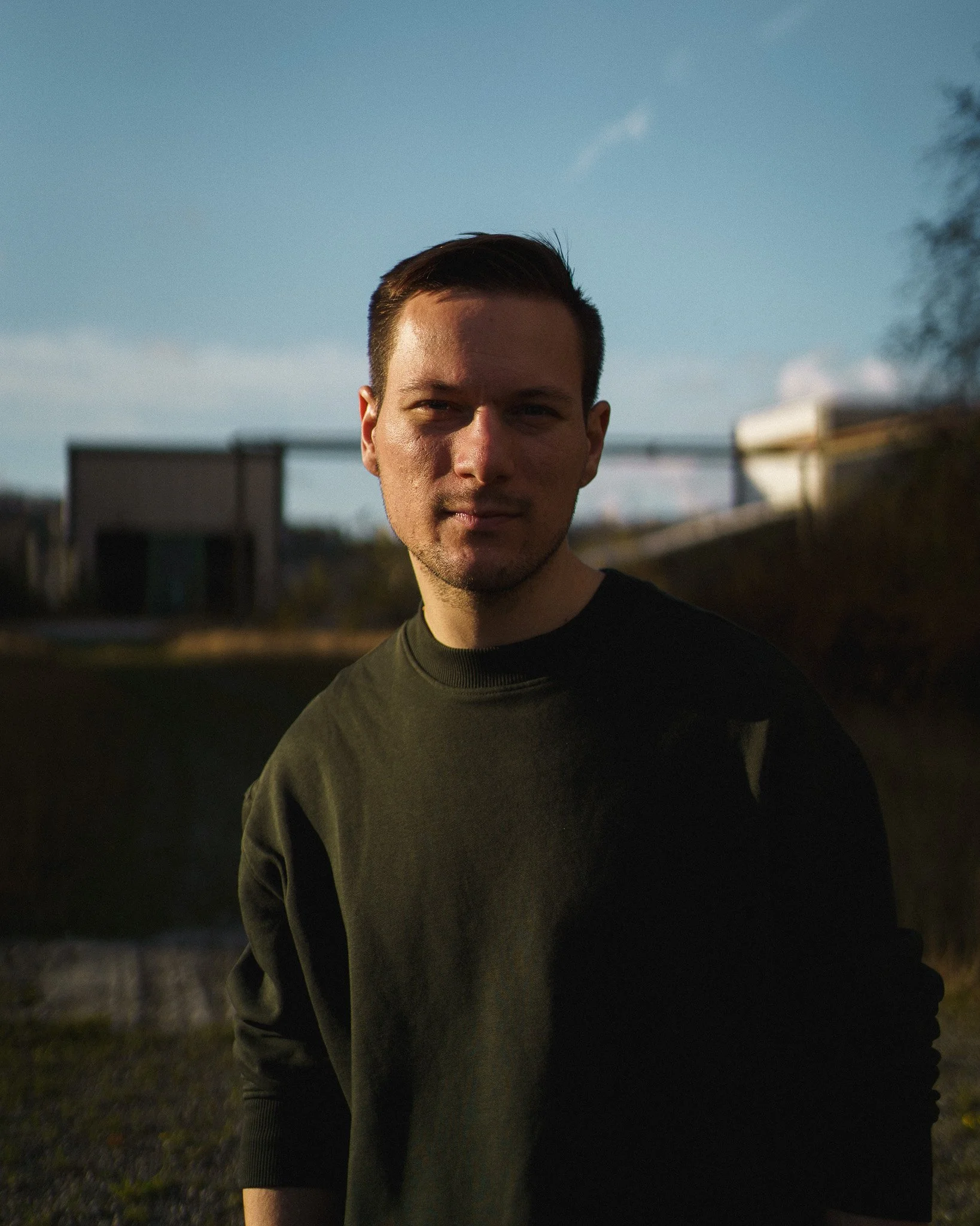 A young man with short dark hair and a slight smile standing outdoors during dusk or sunset, wearing a dark long-sleeve shirt, with a background of blurry houses and trees under a partly cloudy sky.
