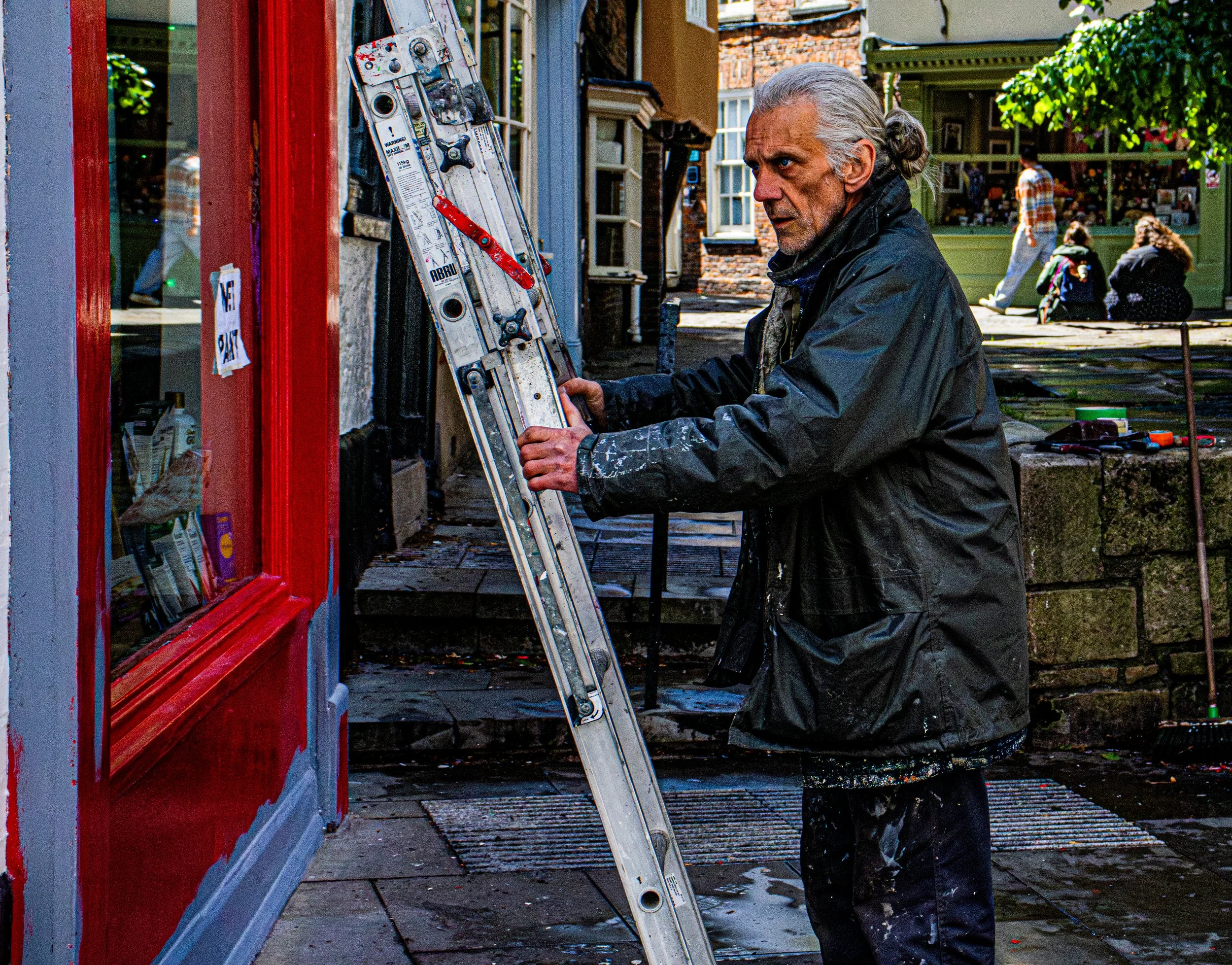 Older man with gray hair wearing a black raincoat working on a bicycle repair or maintenance outside on a city sidewalk, with a red storefront window on the left and people sitting and walking in the background.