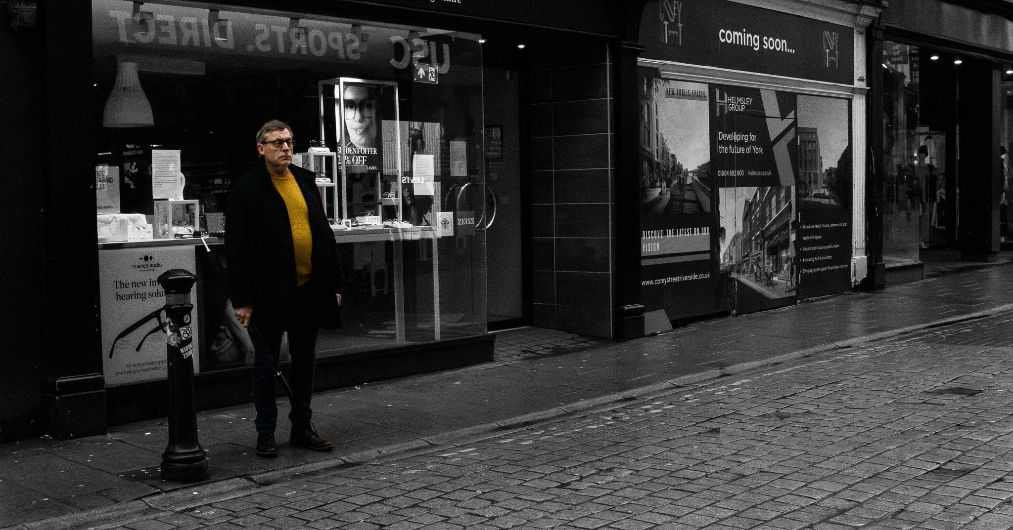 A man standing outside a closed shop on a rainy city street at night. The shop window displays eyeglasses and advertisements. The sidewalk and street are wet, and there are other storefronts and pedestrians in the background.