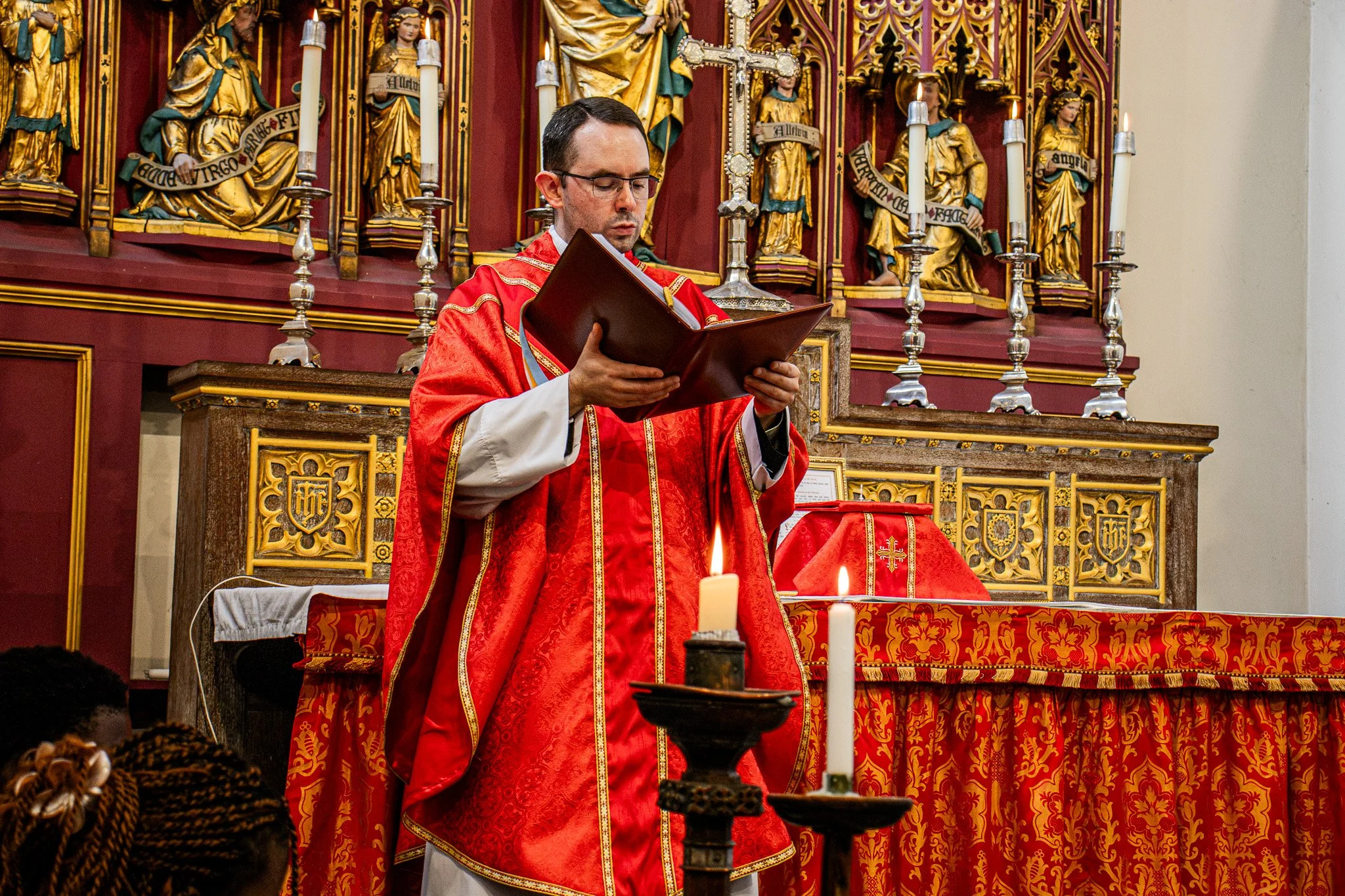 A priest or clergy member in red and white religious robes standing in a church, holding a book, with lit candles, red and gold religious decorations, and statues of saints or religious figures in the background.