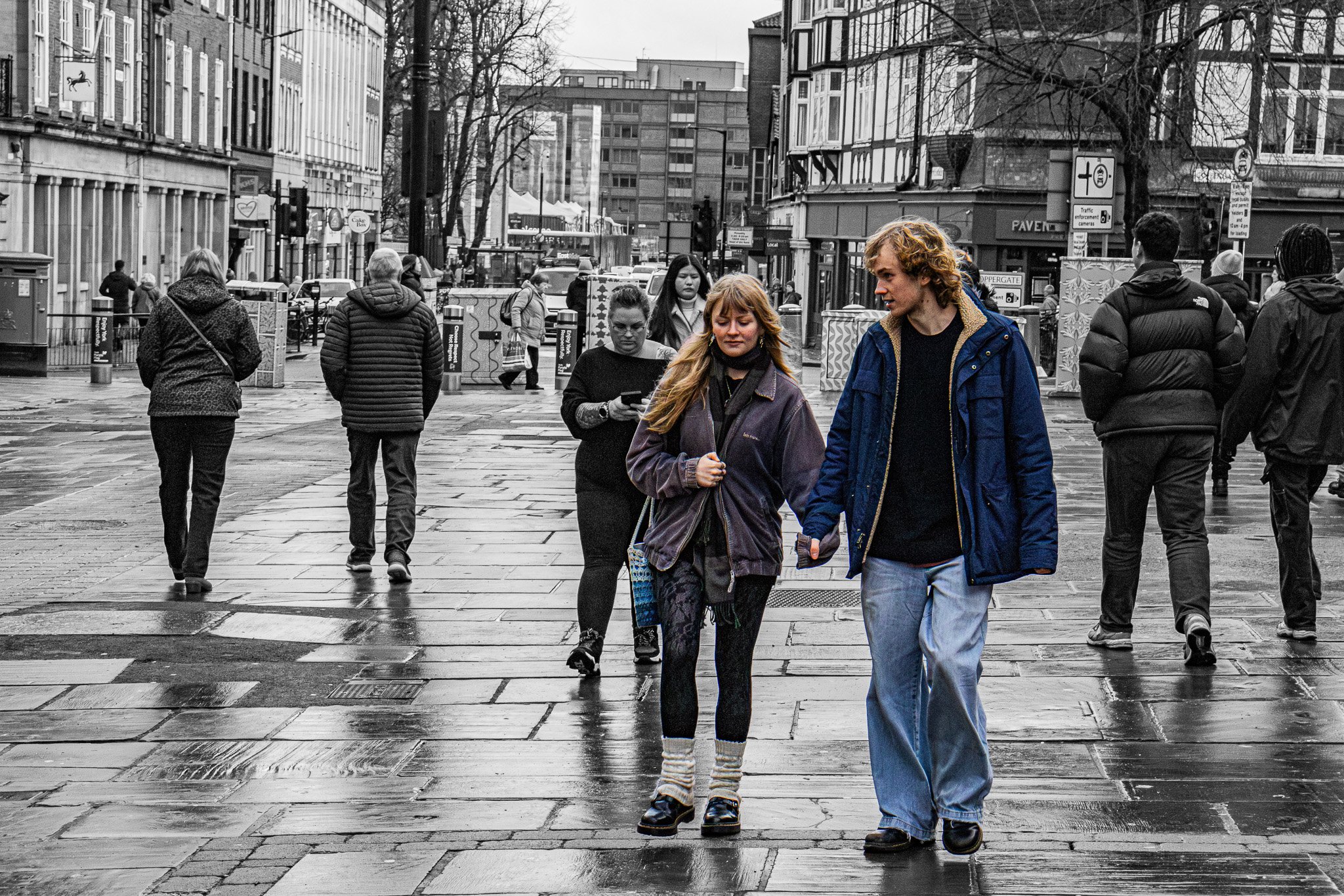 People walking on a wet city street with shops and buildings in the background, some crossing a pedestrian crosswalk.
