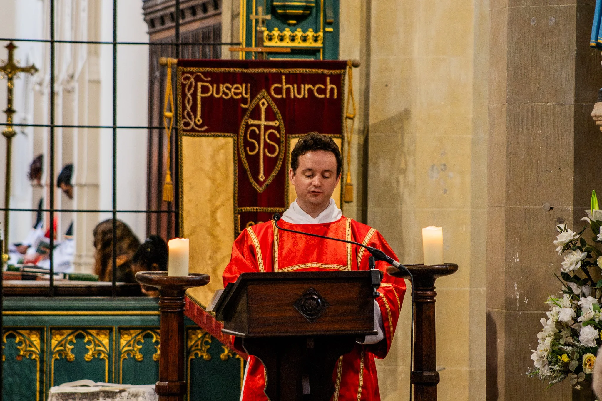 A man in red liturgical vestments standing at a pulpit in a church, with candles on either side, in front of a banner that reads 'Pusey church' with a religious emblem.