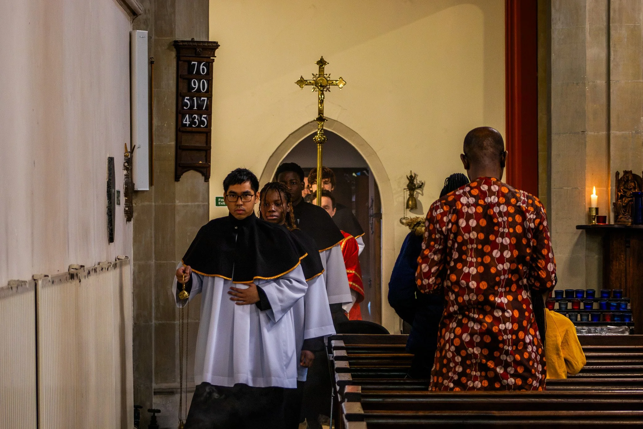 People inside a church, some dressed in religious attire, possibly during a religious ceremony or procession, with a cross and hymn boards visible in the background.