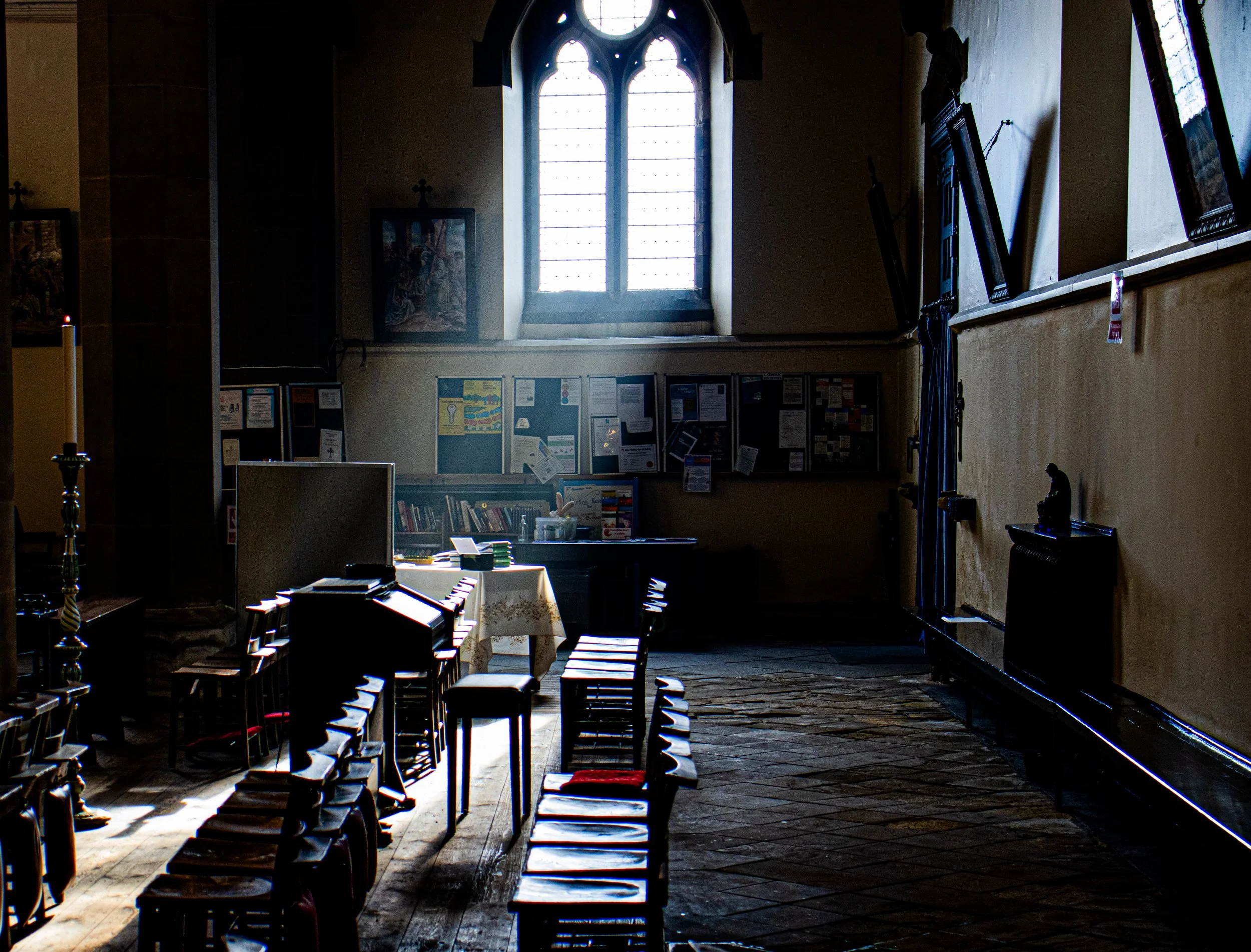 Interior of a church with sunlight coming through a tall arched stained glass window, wooden chairs arranged in rows, and framed religious artwork on the walls.