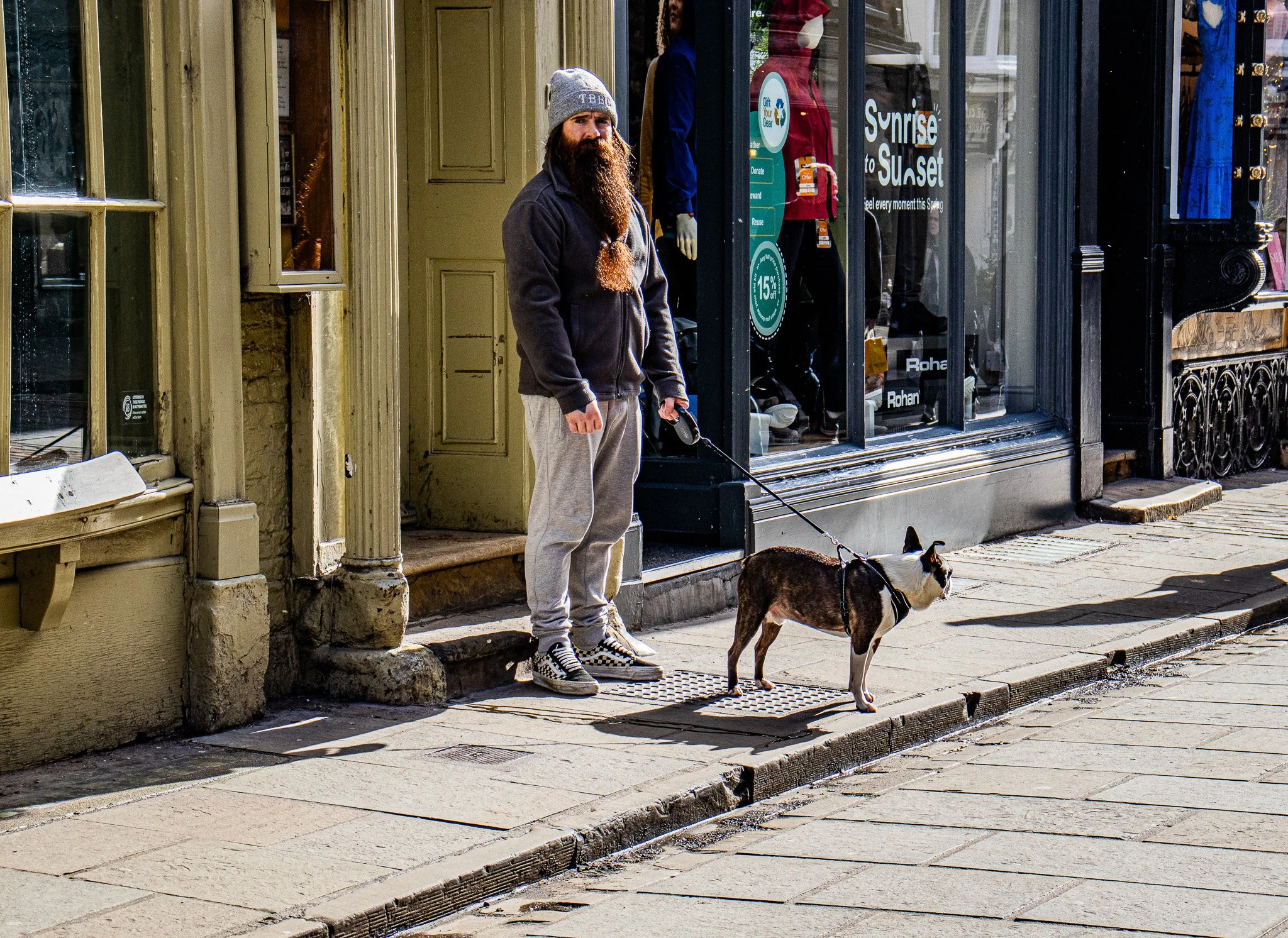 A man with a long beard and long hair wearing a gray beanie, dark jacket, and loose pants standing on the sidewalk while holding a leash attached to a dog. The man is positioned near a storefront window with clothing displays and signs, and there is 