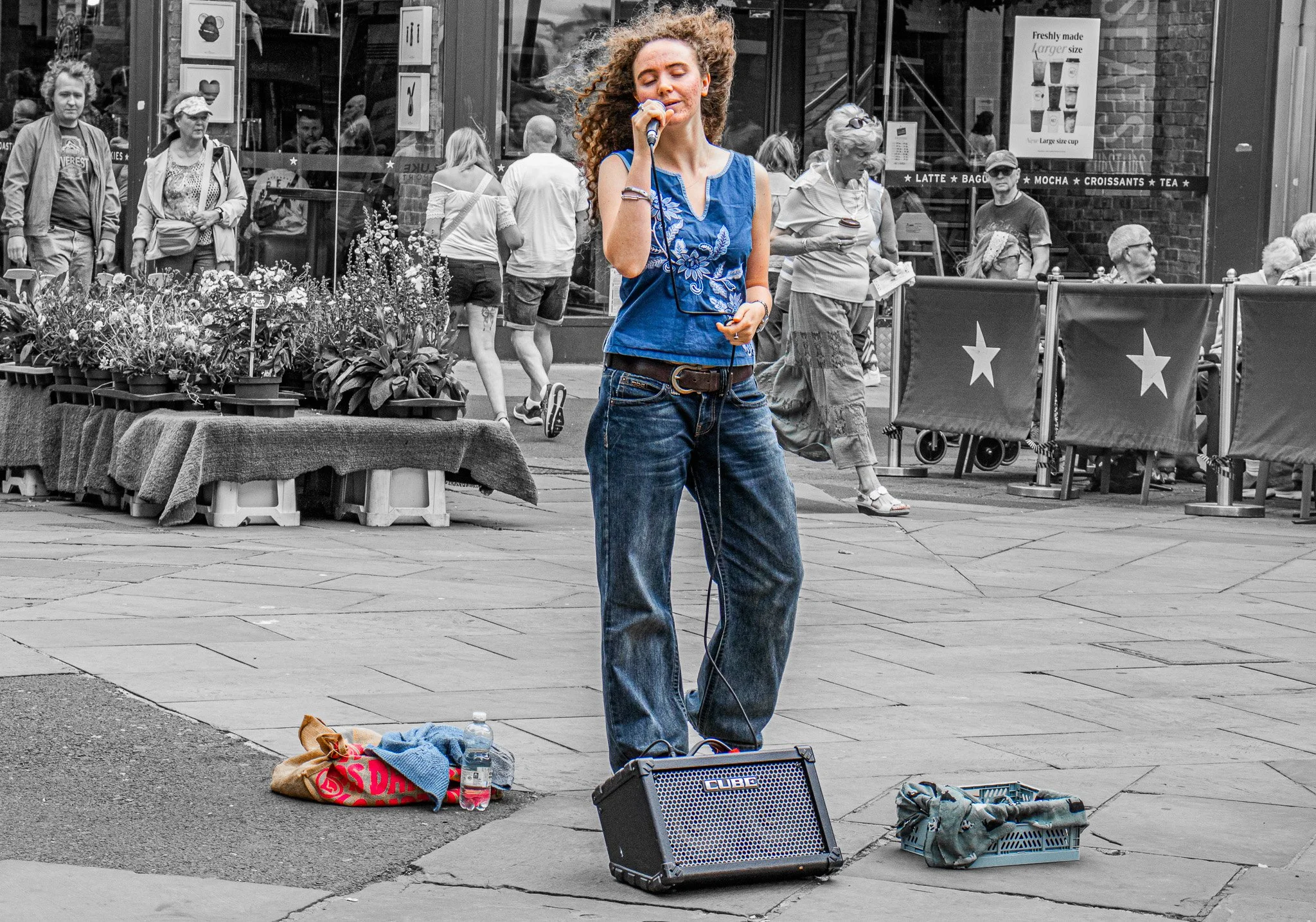 A woman with curly hair singing into a microphone on a city street, with clothes and belongings scattered at her feet, while people pass by in the background.
