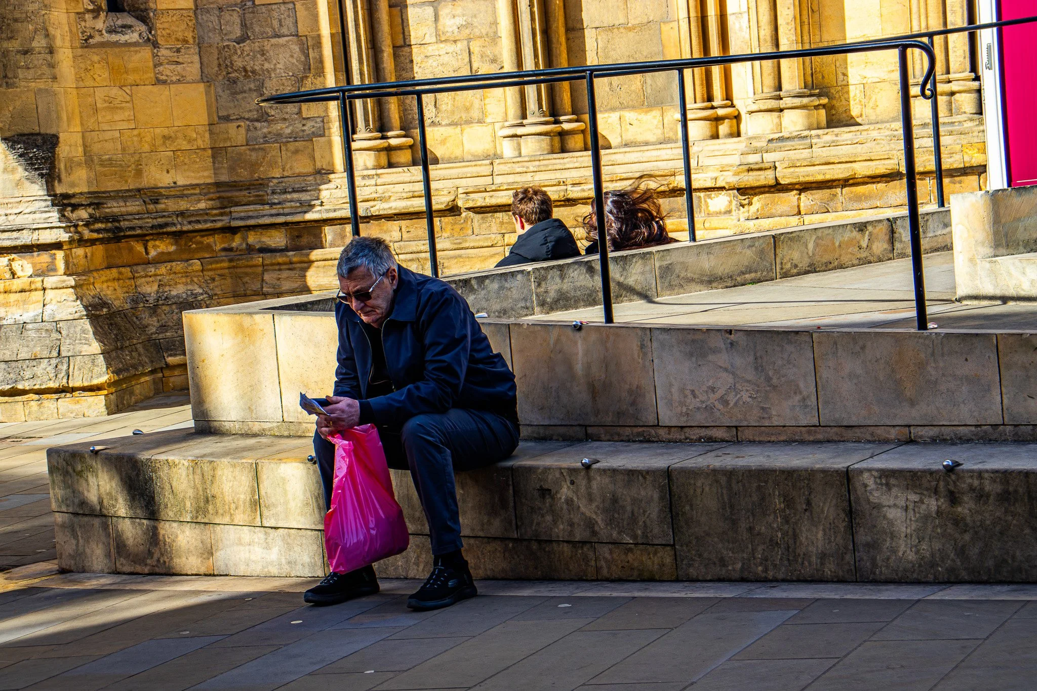 An older man with sunglasses sitting on a stone bench, holding a pink shopping bag and looking at his phone. Behind him, two younger people sit on a higher bench, with their backs turned, near a historic building with stone walls and black railings.