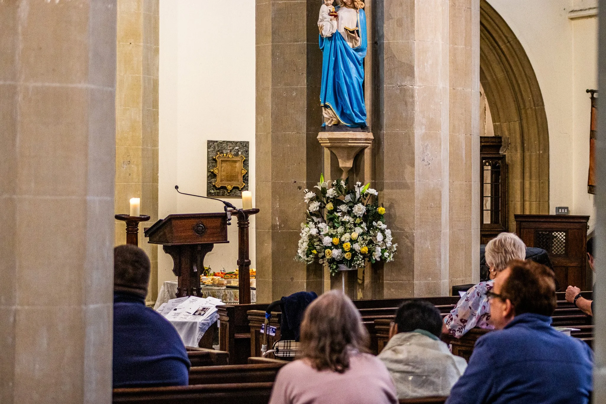 People gather inside a church, sitting on wooden pews, during a religious service. There is a statue of the Virgin Mary dressed in blue and white, placed on a pedestal with flowers below. Candles are lit on stands nearby, and a lectern is visible in the foreground.