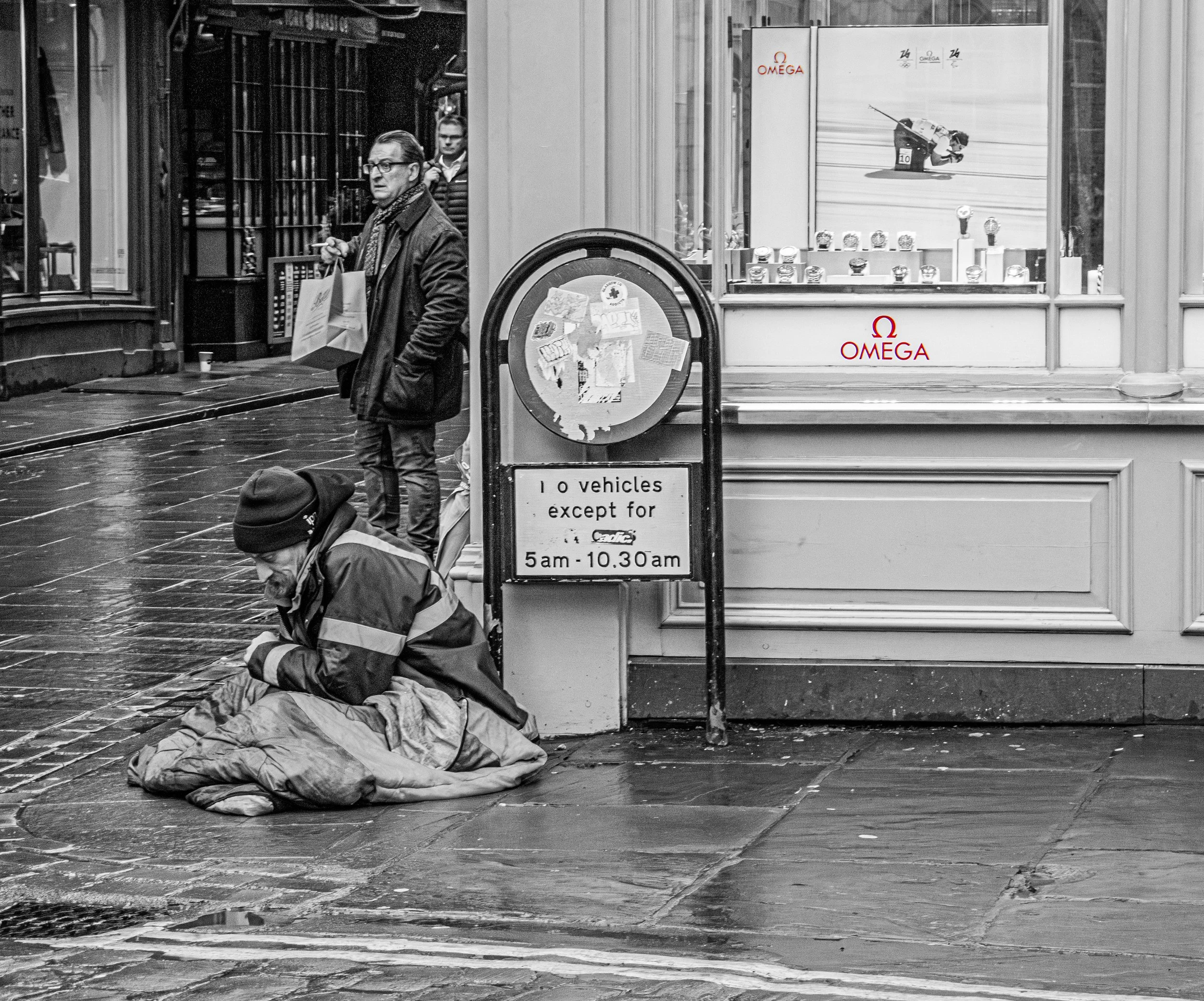 A black and white photo of a street scene featuring a person lying on the cobblestone sidewalk covered with a blanket, a standing man wearing glasses and a scarf holding a shopping bag, and another person standing behind him. The background includes 