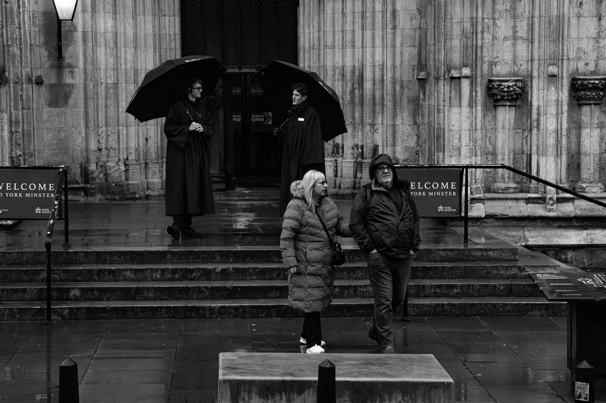 Two people wearing dark coats holding umbrellas, standing and talking to each other on wet steps outside a stone building with columns, while two others walk past in the foreground.