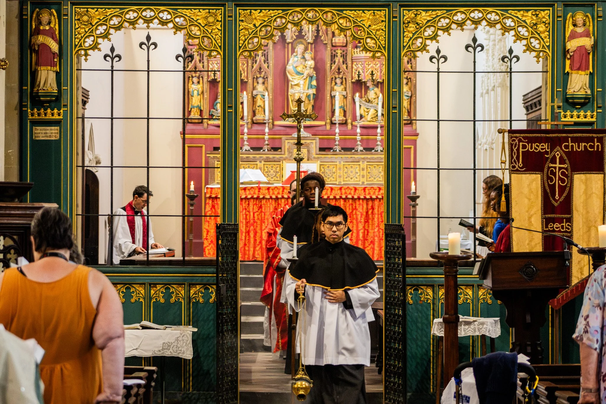 Inside Pusey Church during a service, a young man dressed in a white and black choir robe stands in front of the altar holding a staff. The altar features candles and a large religious painting of Madonna and Child behind ornate gold and woodwork. People are seated and standing, some reading or listening, with the church's interior visible.