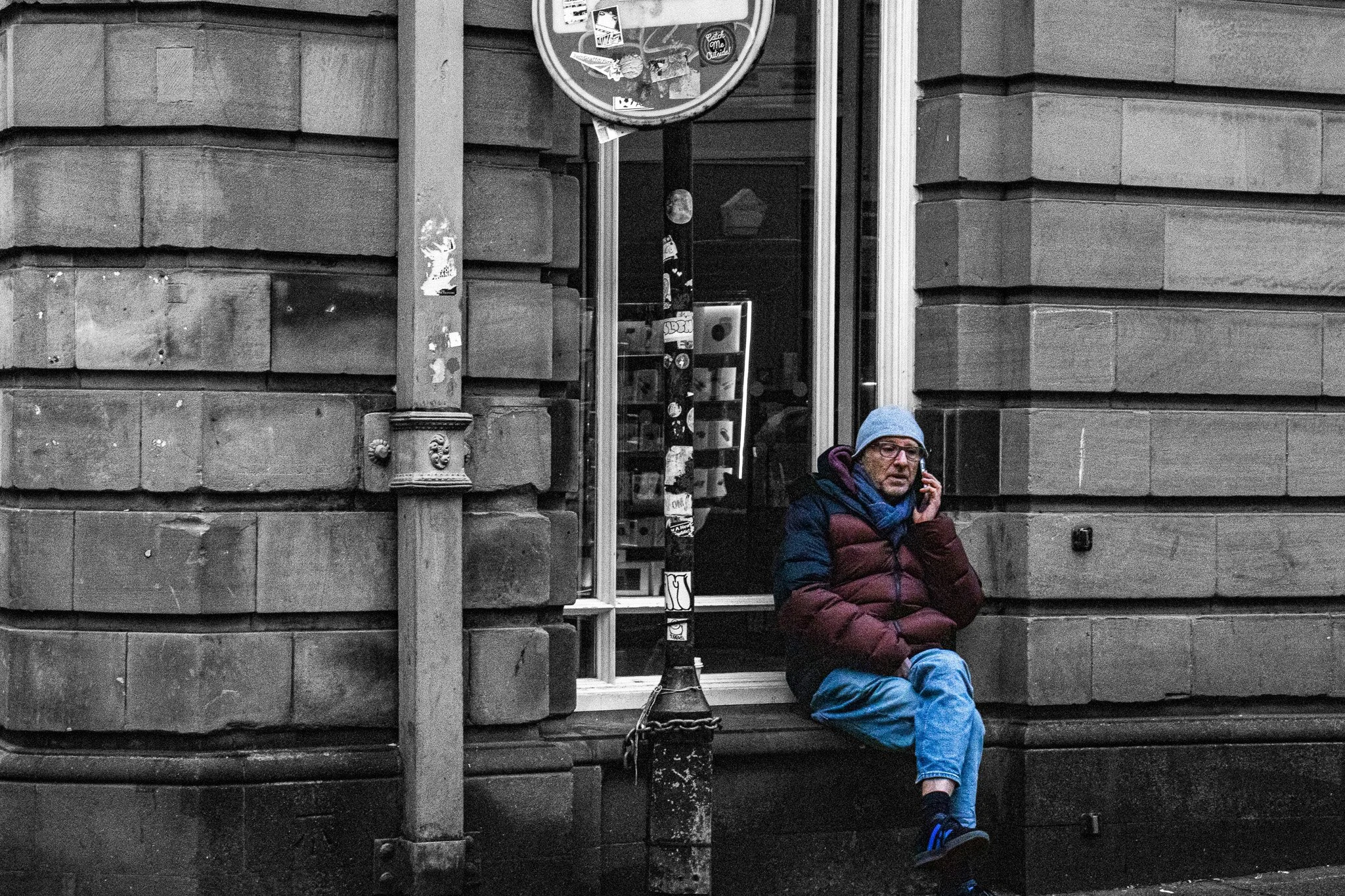 A man in a blue hoodie and glasses sitting on the sidewalk outside a building, talking on a cellphone.