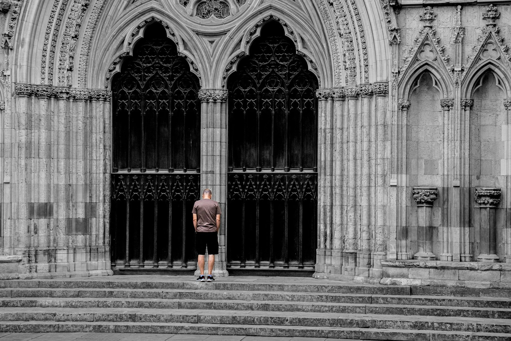 A person stands at the bottom of stone steps, facing a large gothic-style cathedral door with intricate carvings and architectural details, in black and white.