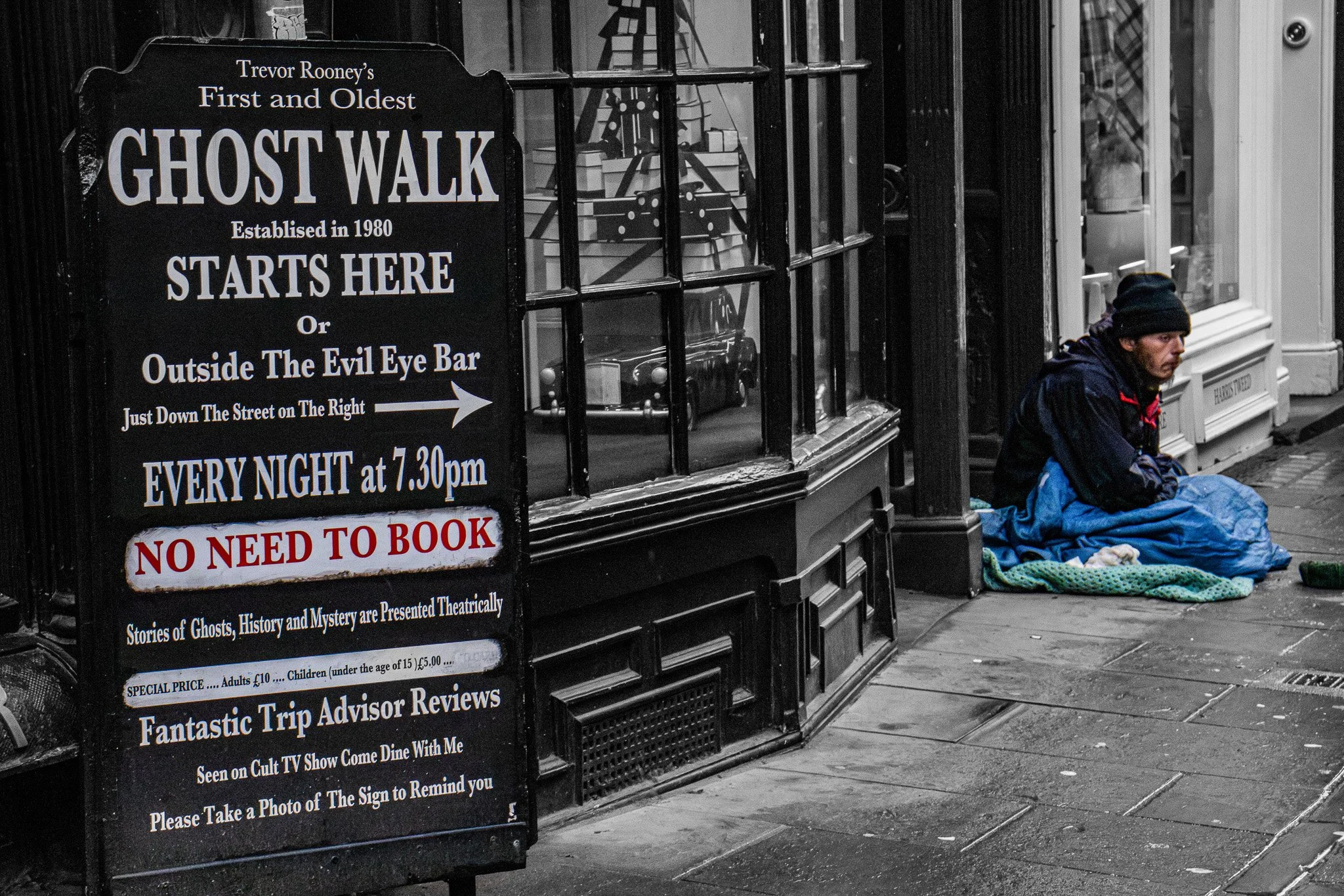 A person sitting on the sidewalk with a blanket, next to a sign that reads 'Trevor Rooney's First and Oldest Ghost Walk, Established in 1980, Starts Here, or Outside The Evil Eye Bar, Just Down The Street on The Right, Every Night at 7:30 pm, No Need