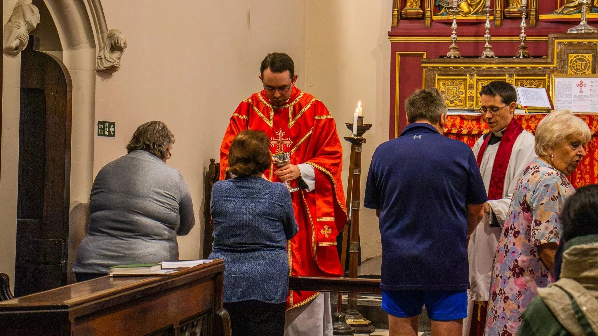 A priest in red robes performing a religious ritual with a congregation of people gathered around inside a church.